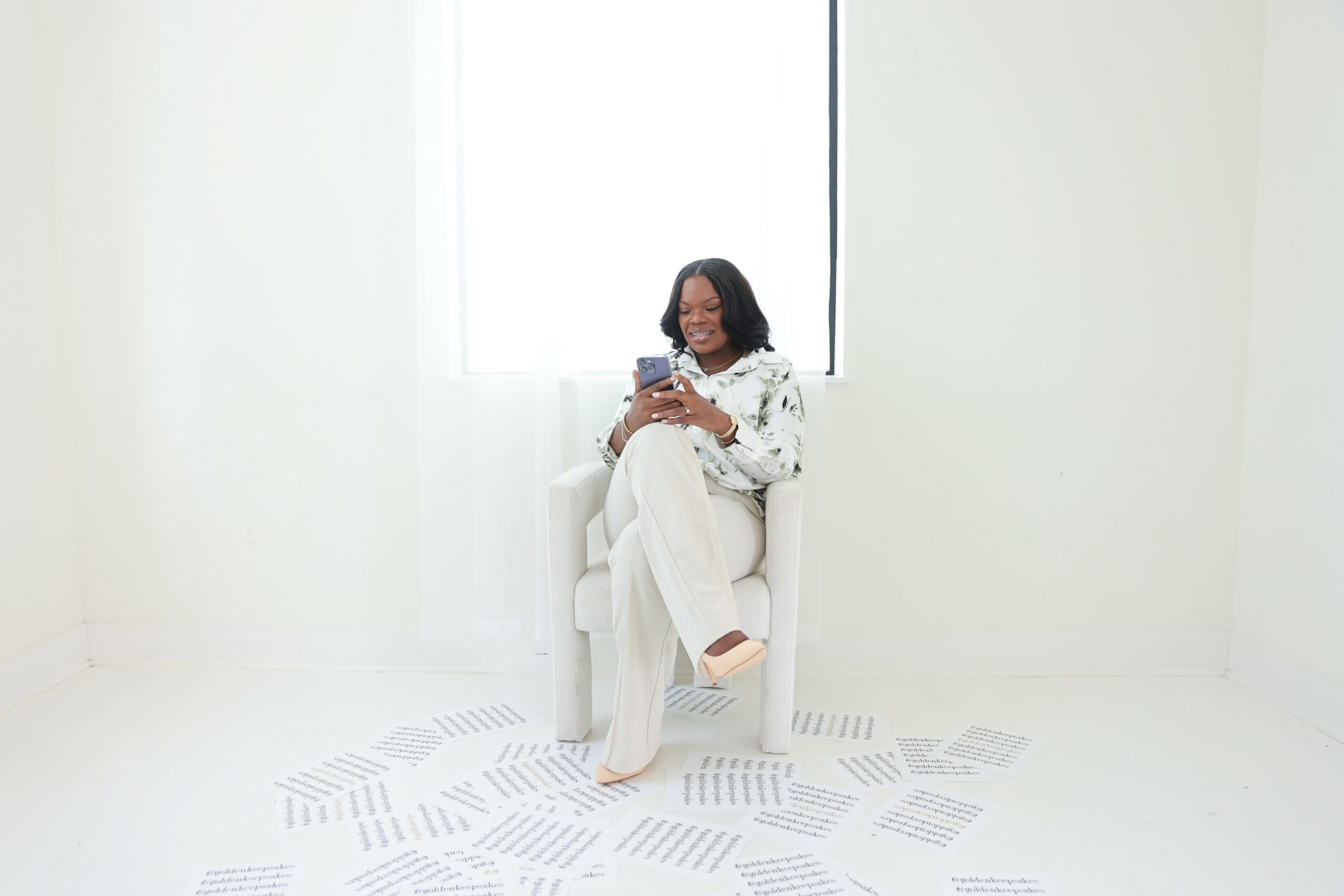 Woman sitting on a white chair in a bright room, holding a phone, with papers scattered on the floor.