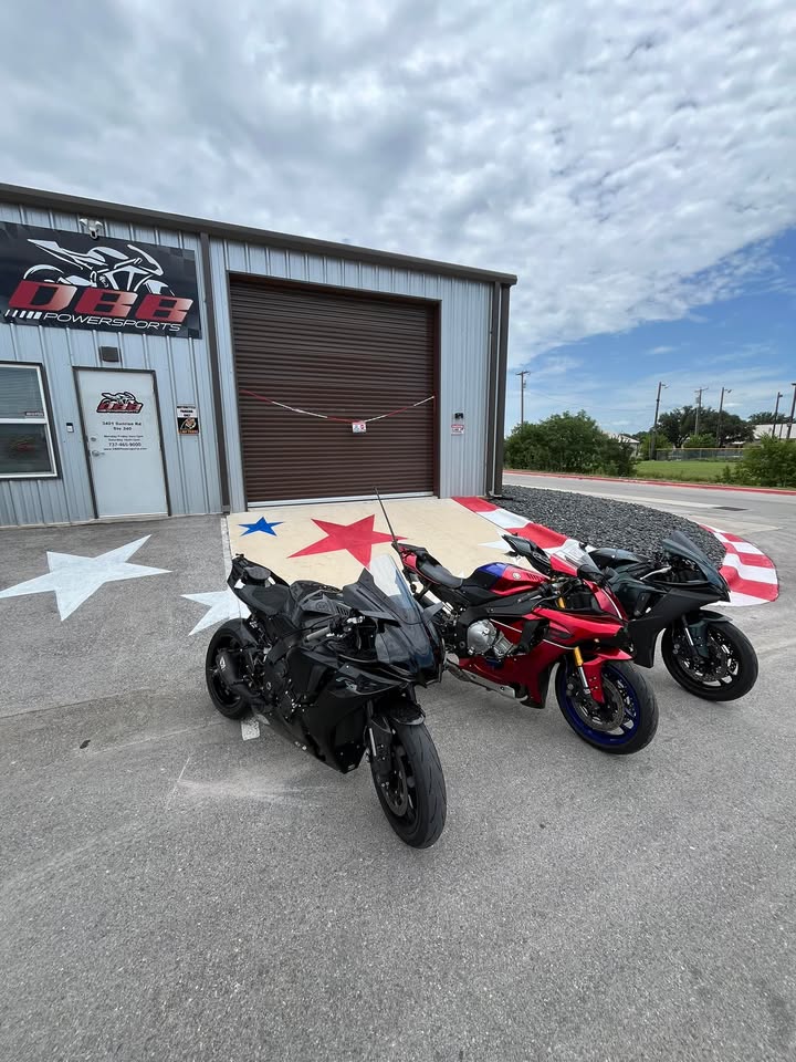 Three motorcycles parked outside a building with a large garage door, stars painted on the ground, and a cloudy sky.