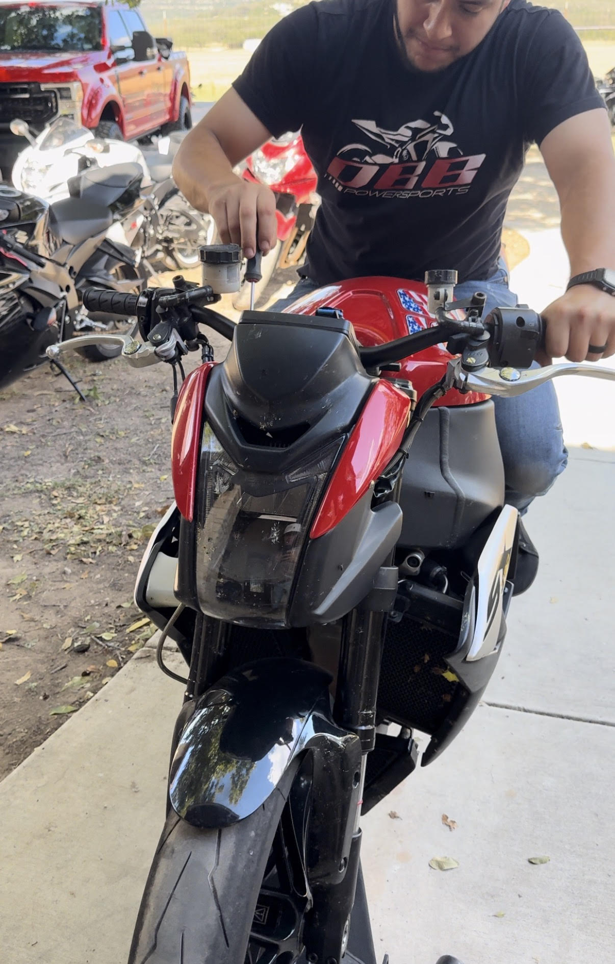 Person standing next to a red and black motorcycle outdoors, with other vehicles in the background.