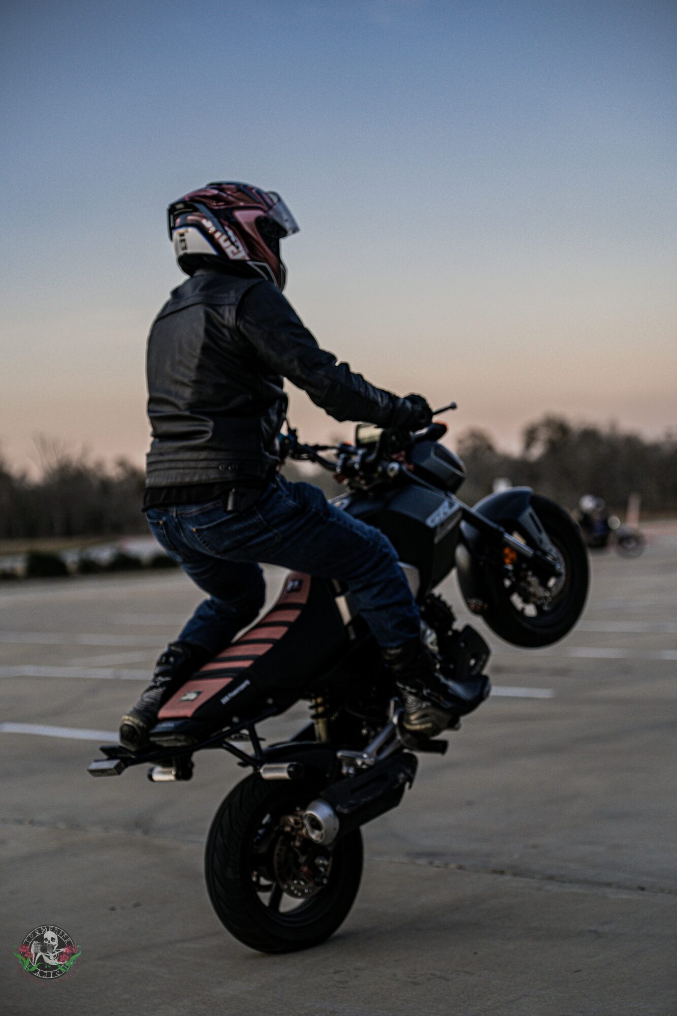Person performing a wheelie on a motorcycle in an open area during dusk, wearing a helmet and protective gear.