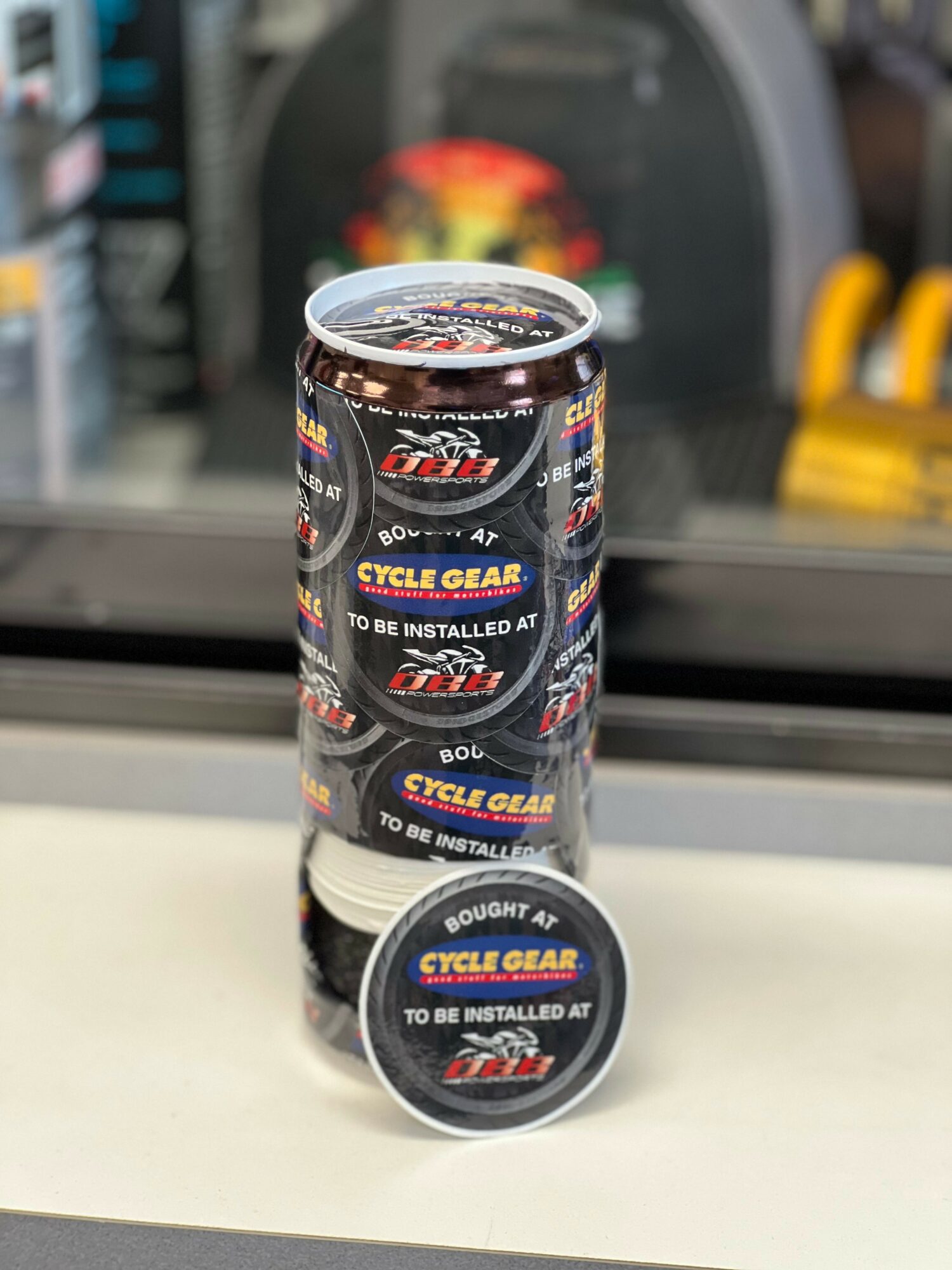 Container of cycle gear lubricant with a cap, placed on a white surface, background shows a blurred store setting.