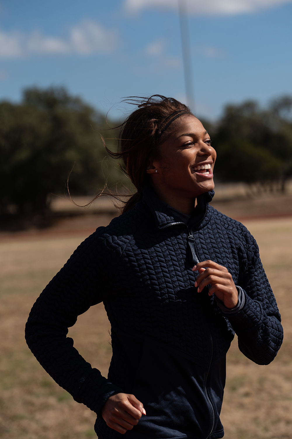 Woman smiling outdoors, wearing a dark jacket, with trees and sky in the background.