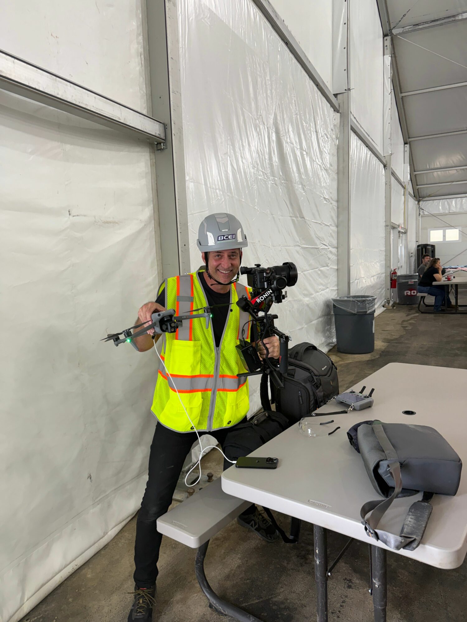 Person in high-visibility jacket and helmet holding a tool, standing next to a white table with a chair, in an industrial setting.