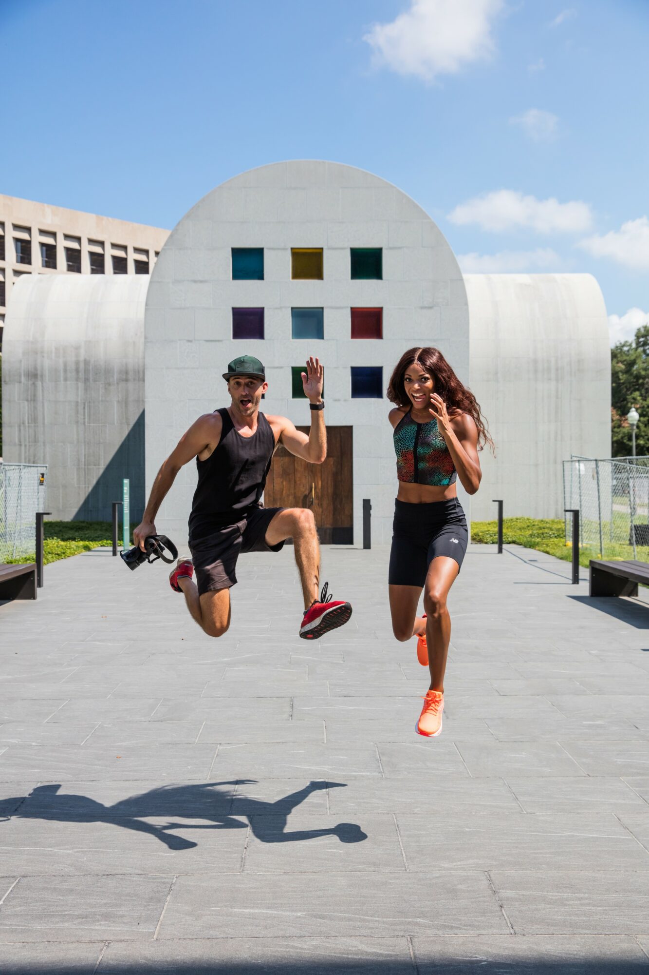 Two people jumping in front of a modern building with a circular window and colorful squares, under a blue sky.