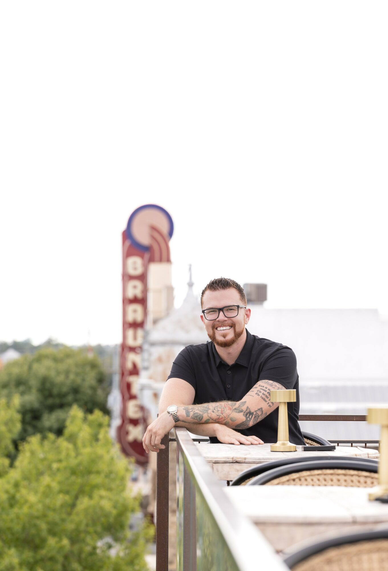 Man with glasses and tattoos leaning on a rooftop railing, smiling, with a cityscape and a vertical sign in the background.