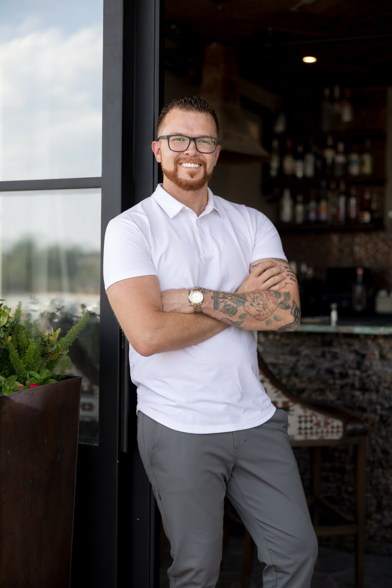 Man with glasses, beard, and tattoos, smiling with arms crossed, standing indoors near a window.
