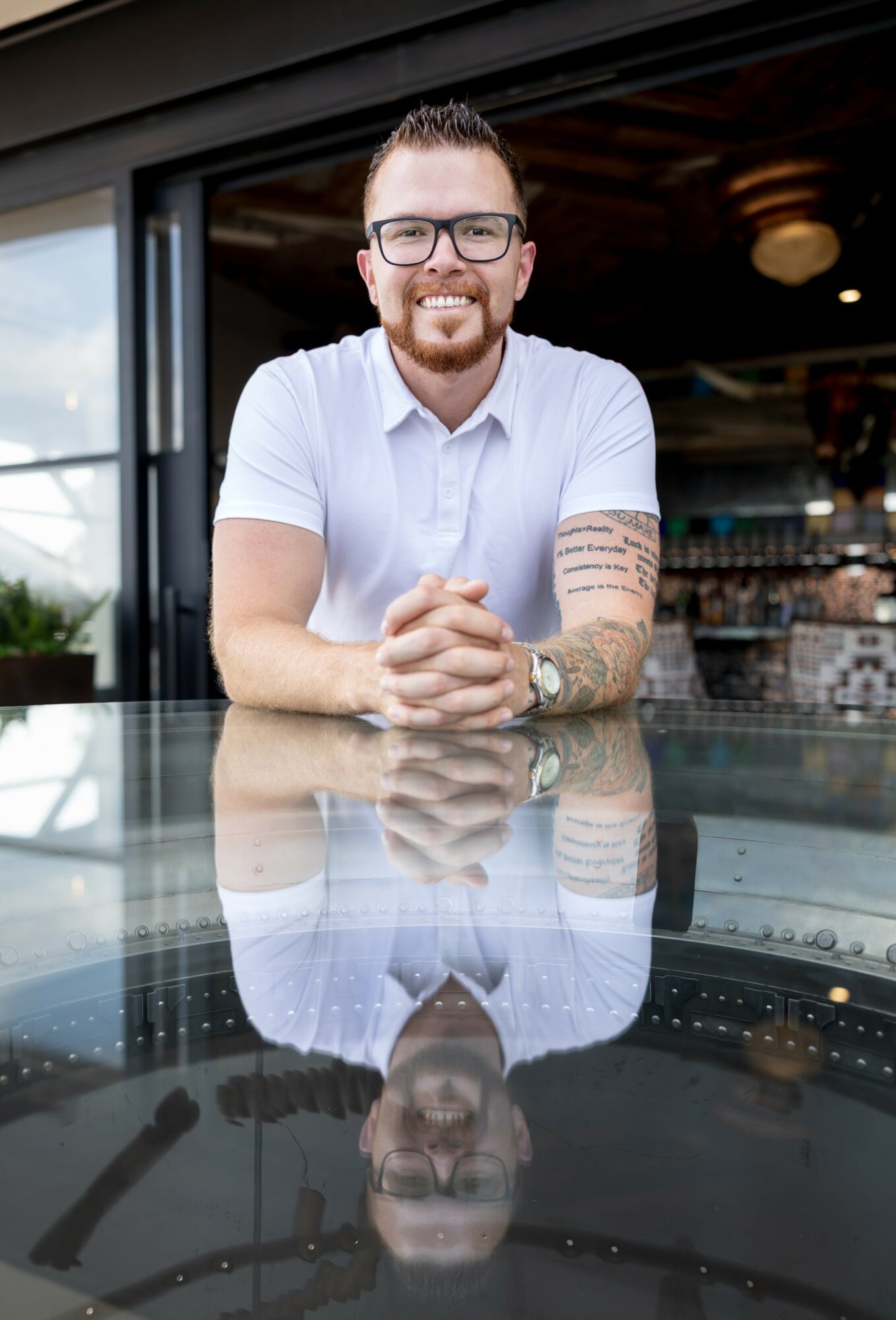 Smiling man with glasses and tattoos, sitting at a reflective table outdoors, with a bar and ceiling fan in background.
