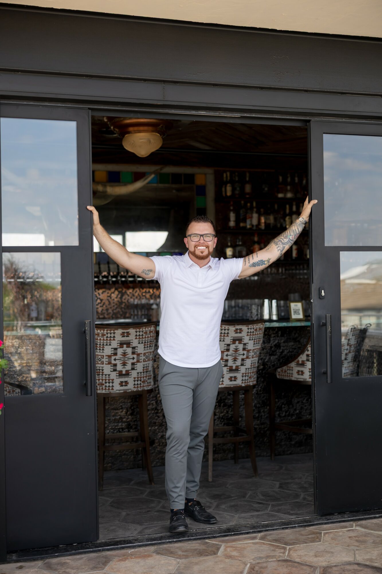 Man with glasses and tattoos stands in doorway with arms outstretched, smiling, inside a bar or cafe.
