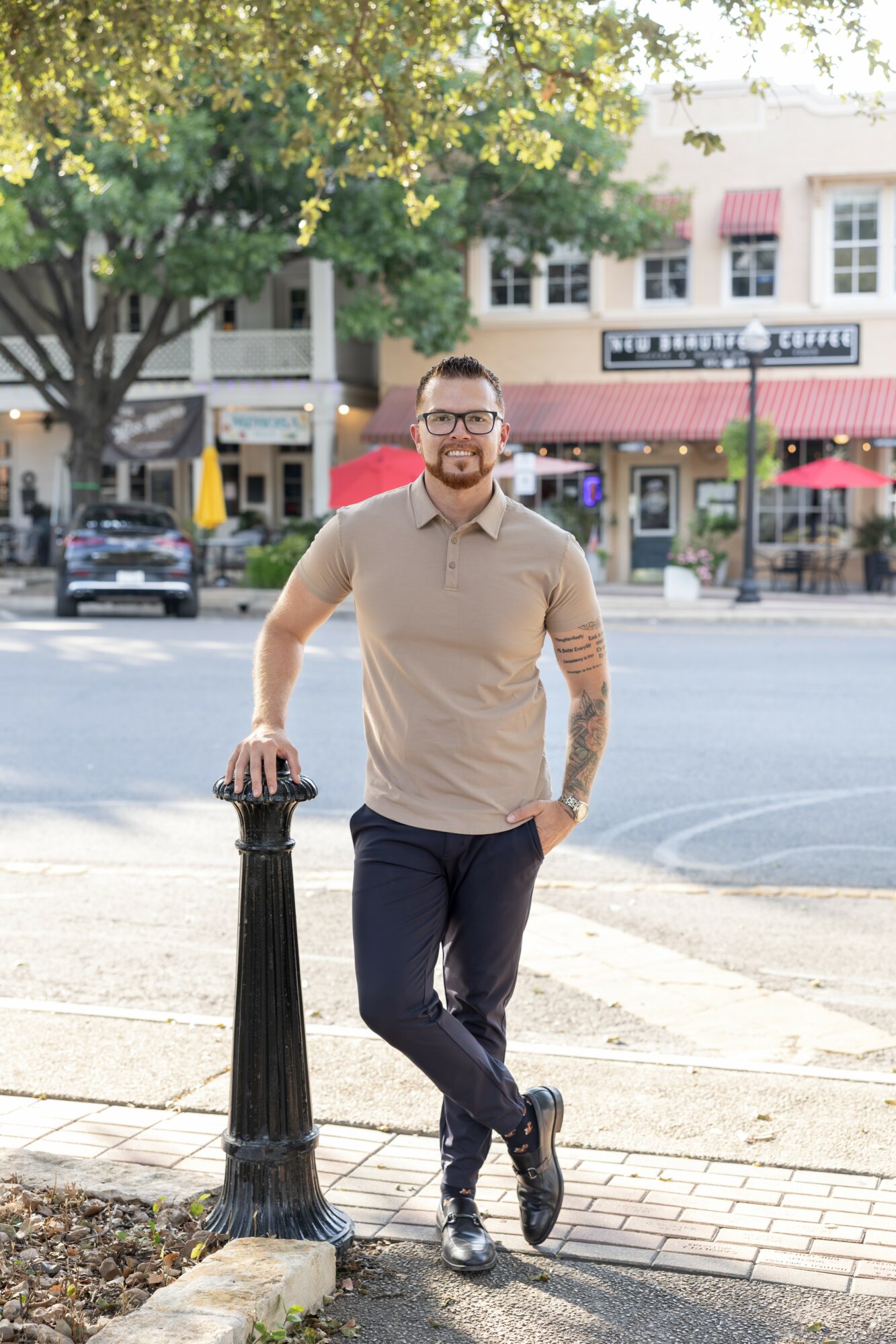 Man standing outdoors on sidewalk, leaning on a black post, with trees and buildings in the background.