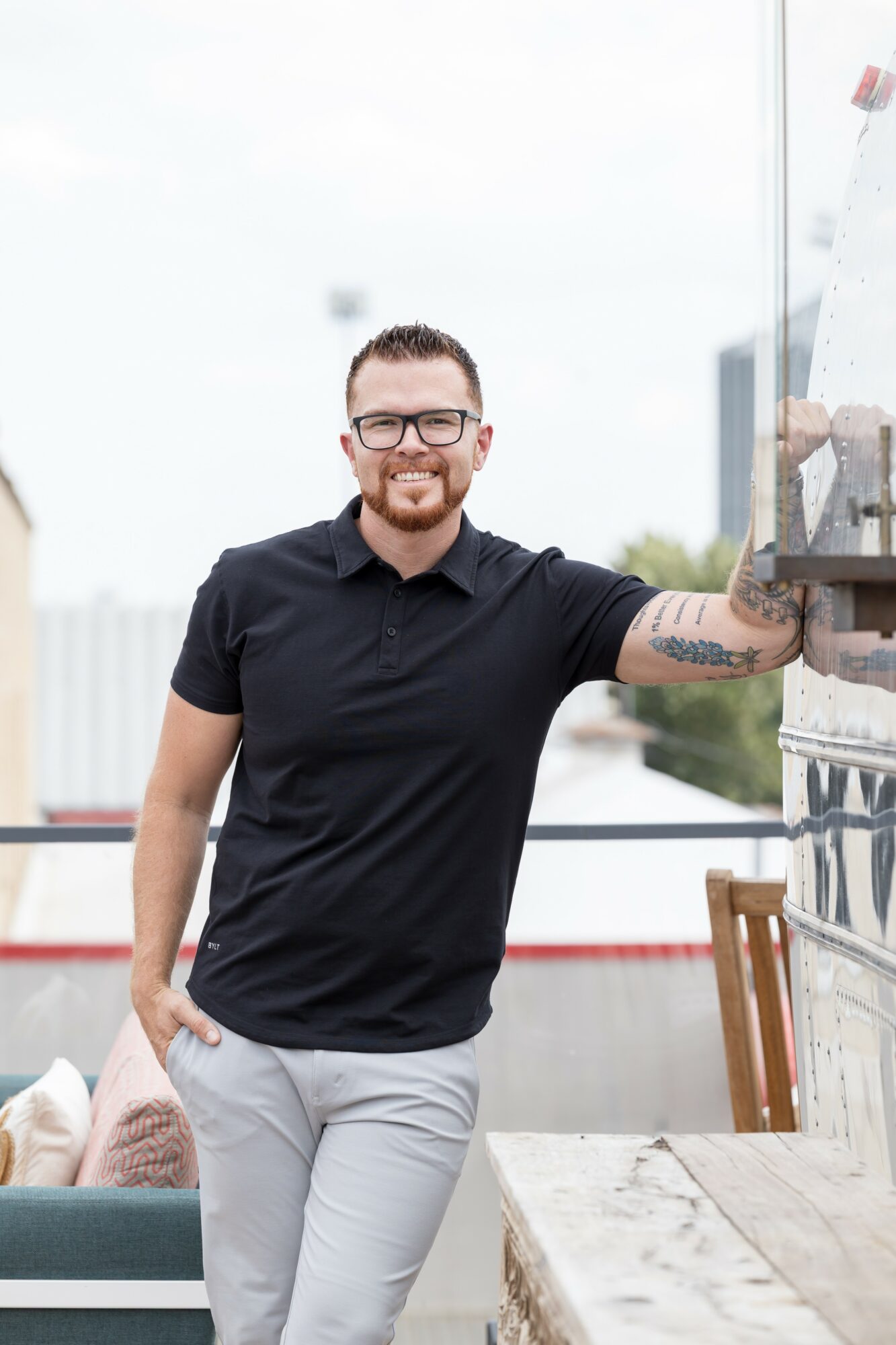 Man with glasses and beard smiling, wearing a black shirt and light pants, standing indoors with city view behind him.