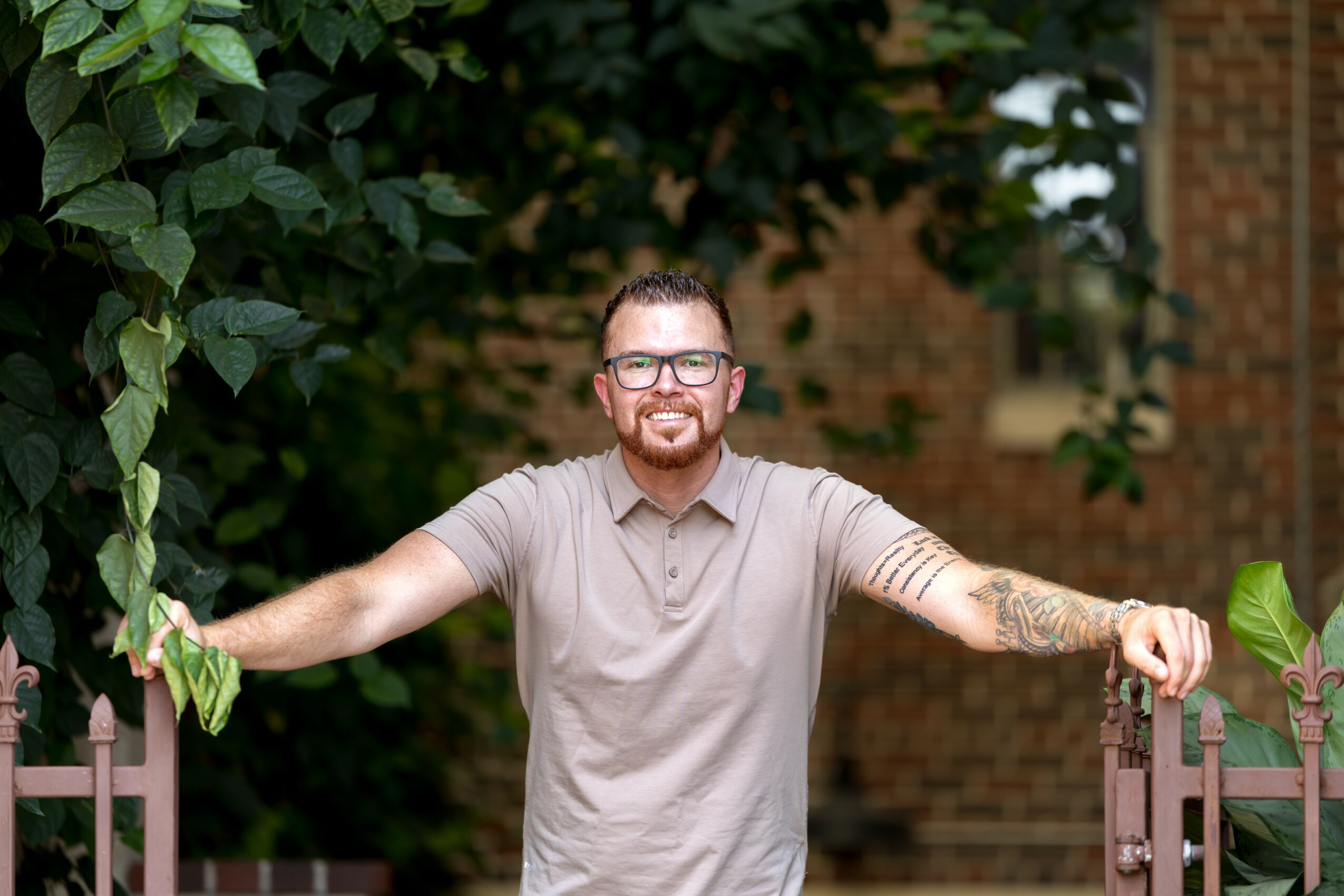 Smiling man with glasses and tattoos extends arms, standing outdoors near a fence and leafy trees.