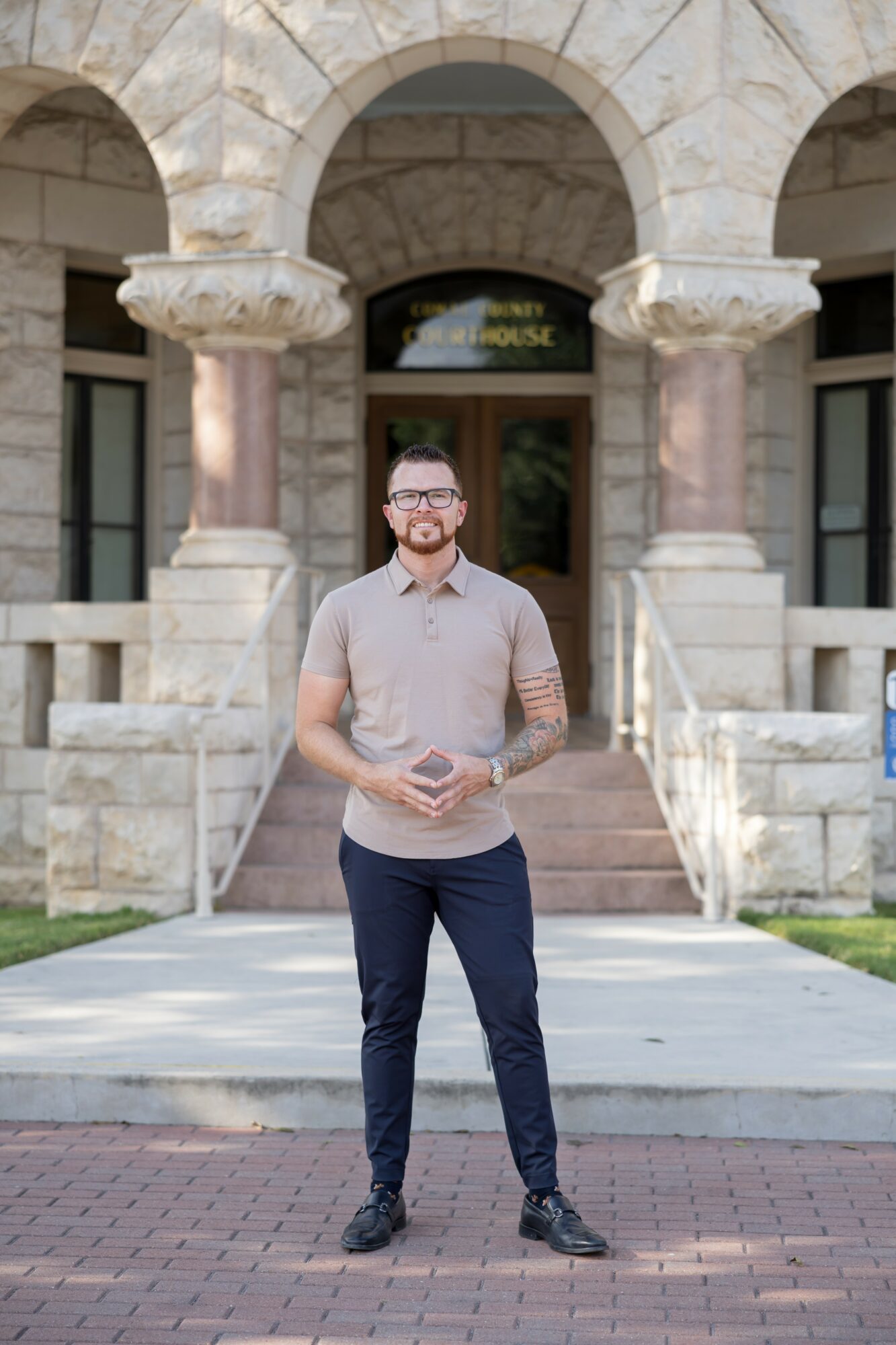A man with glasses and a beard stands outside a stone building with arches, holding a book, wearing a light shirt and dark pants.