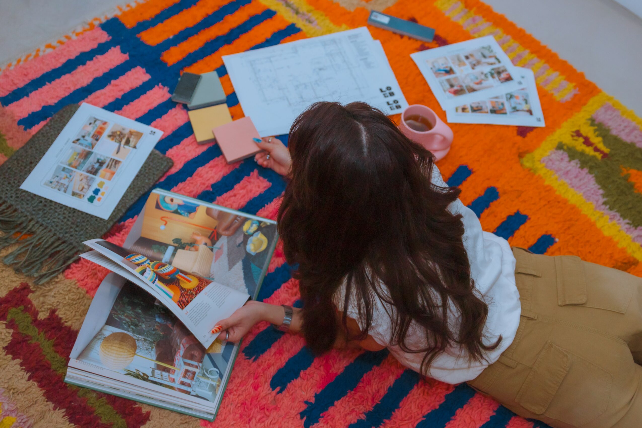 Child with long hair lying on colorful rug, looking at open books and papers, surrounded by magazines and stationery.