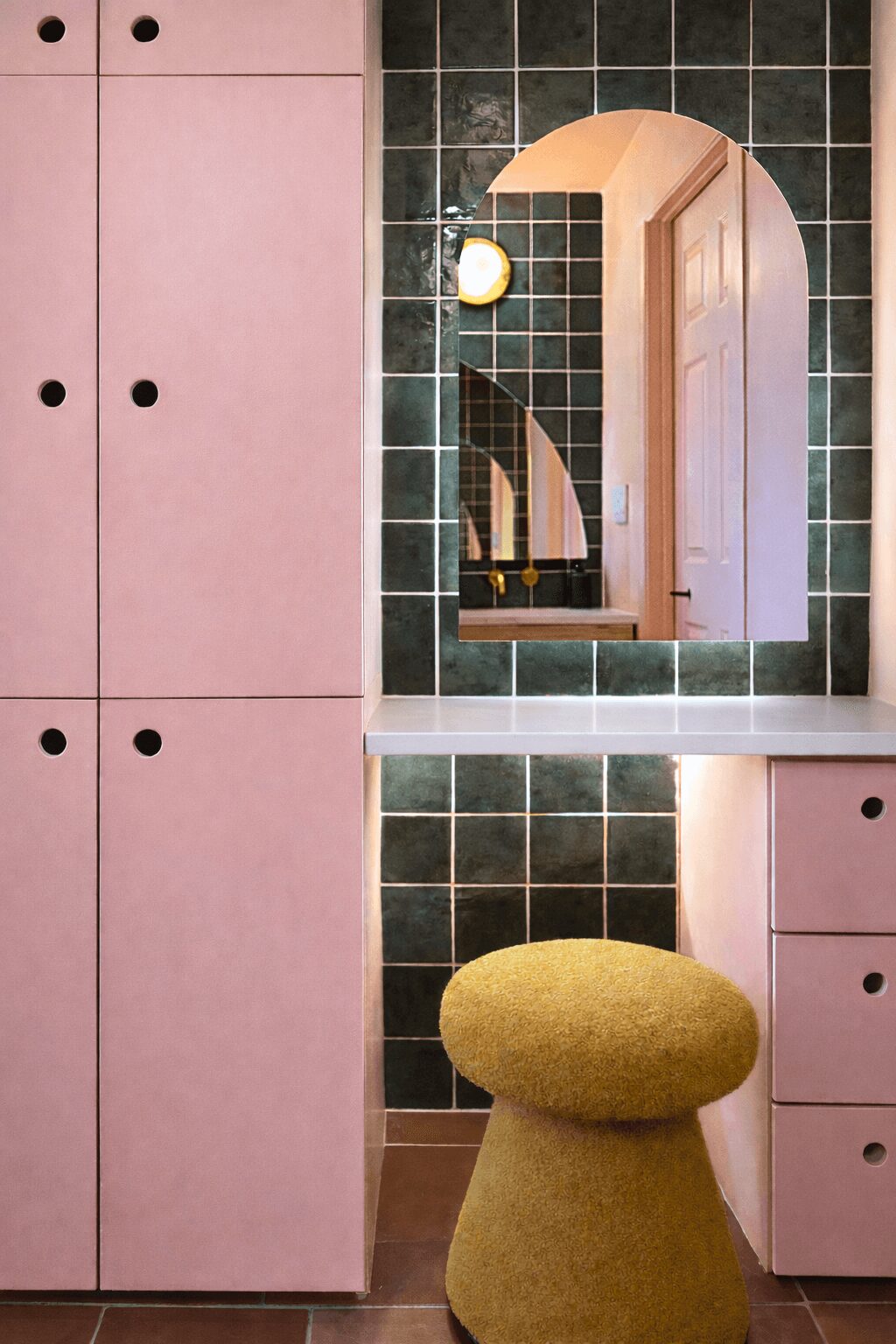 Pink cabinet, green tiled wall, mirror, yellow stool, and a door reflected in the mirror.