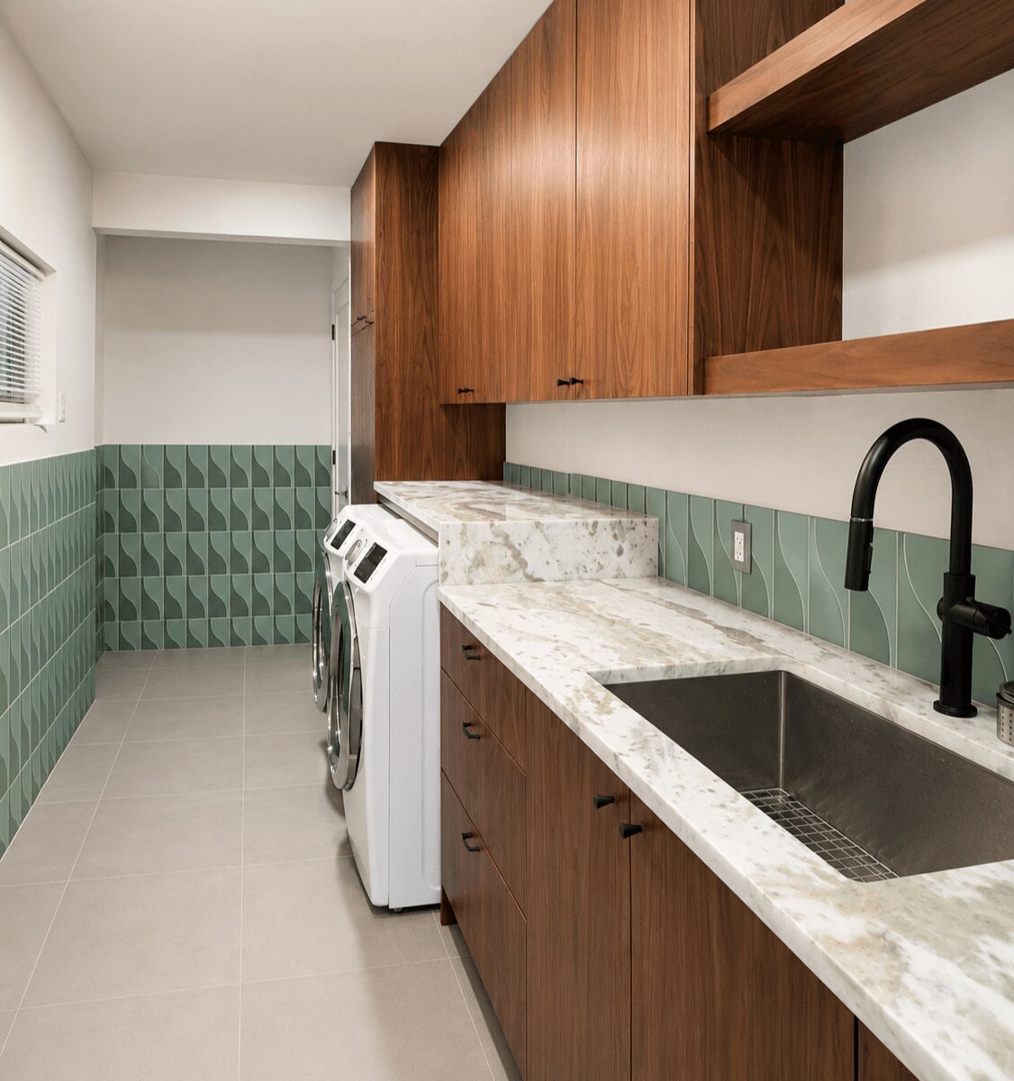 Laundry room with wooden cabinets, marble countertop, sink, and washing machine, green tiled wall, and window with blinds.