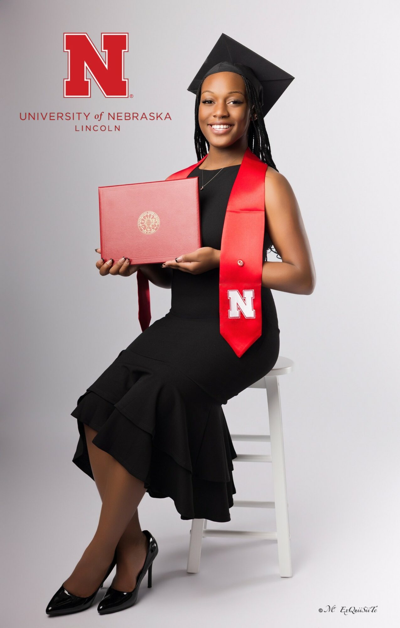 Young woman in graduation cap and gown holding a diploma, sitting on a white chair, with Nebraska University logo in background.