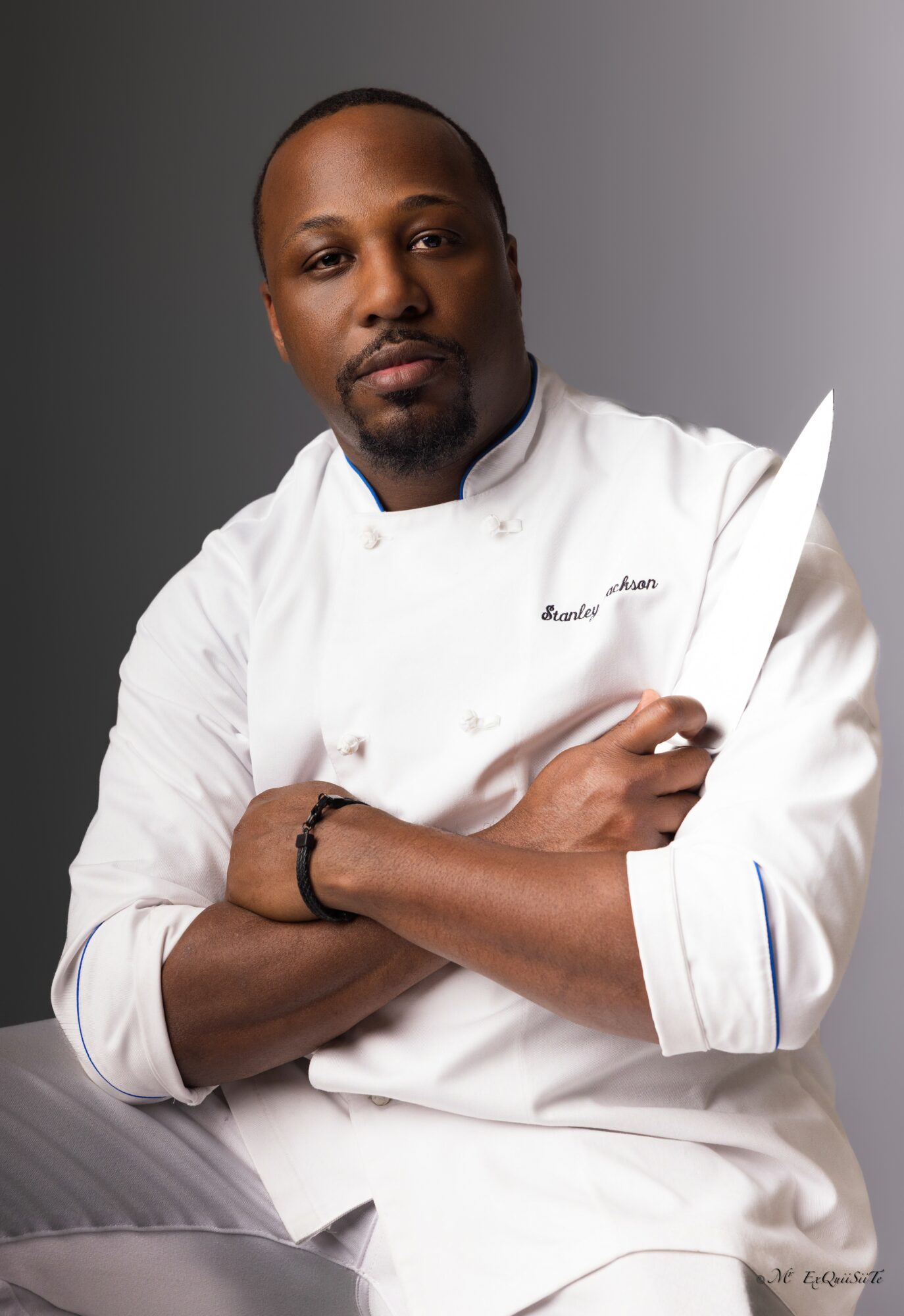 Male chef in white uniform holding a large knife, arms crossed, against a gray background.