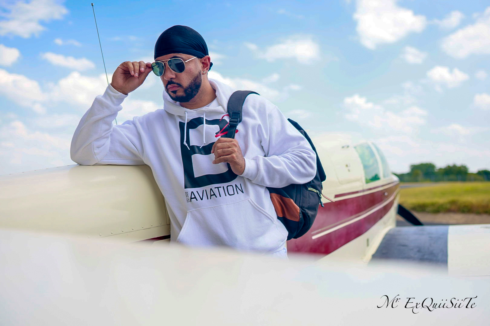Man with sunglasses and a backpack standing next to a boat on a sunny day with clouds in the sky.