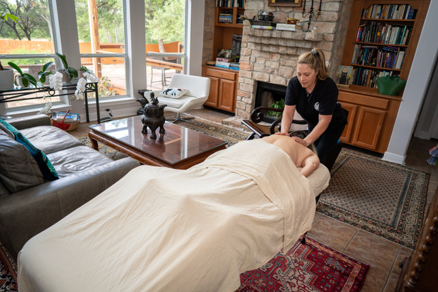 Person lying on a massage table covered with a white sheet, receiving a massage from a therapist in a living room.