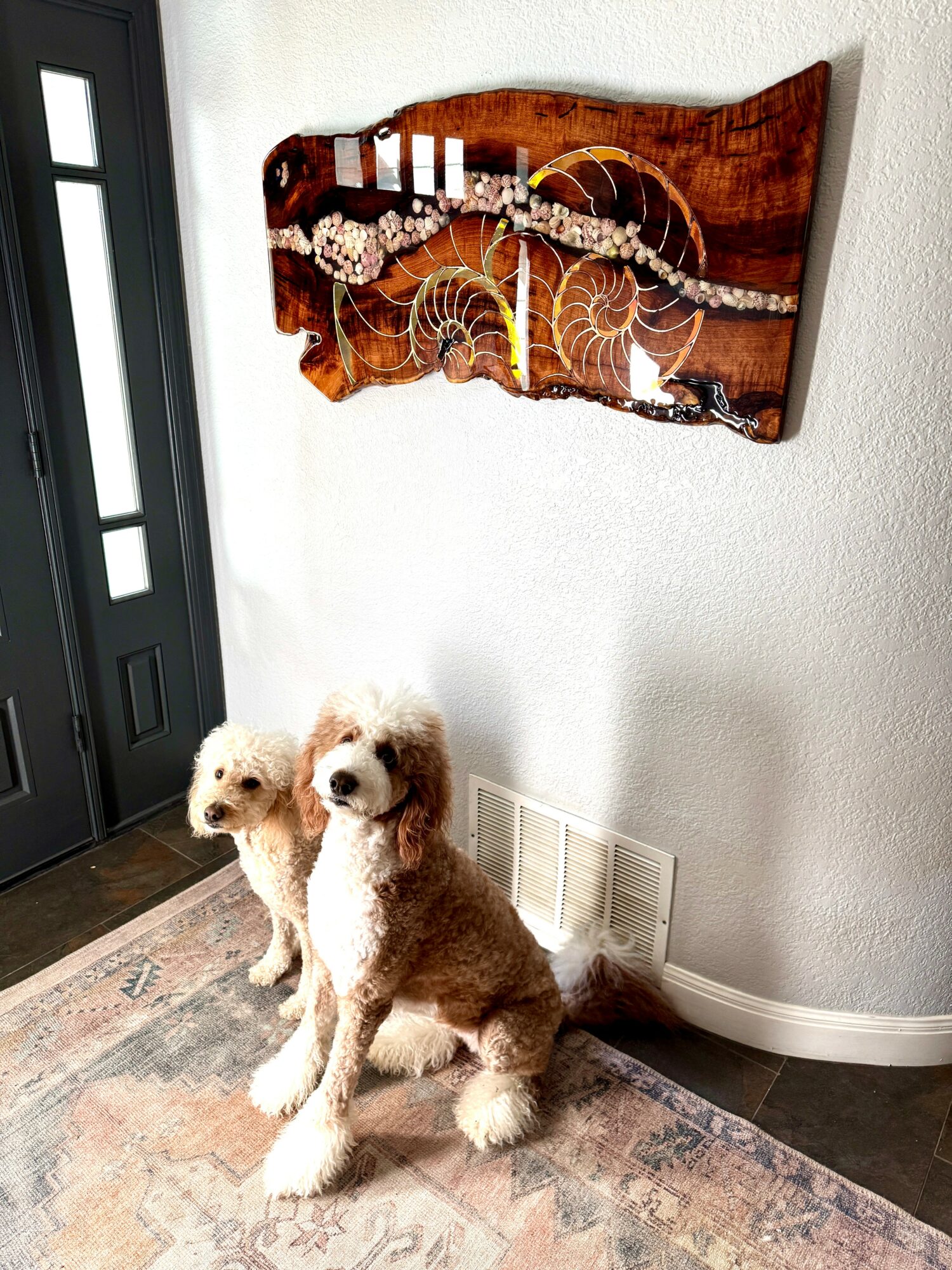 Two dogs sitting on a rug near a wall with a wooden art piece above them, next to a door.