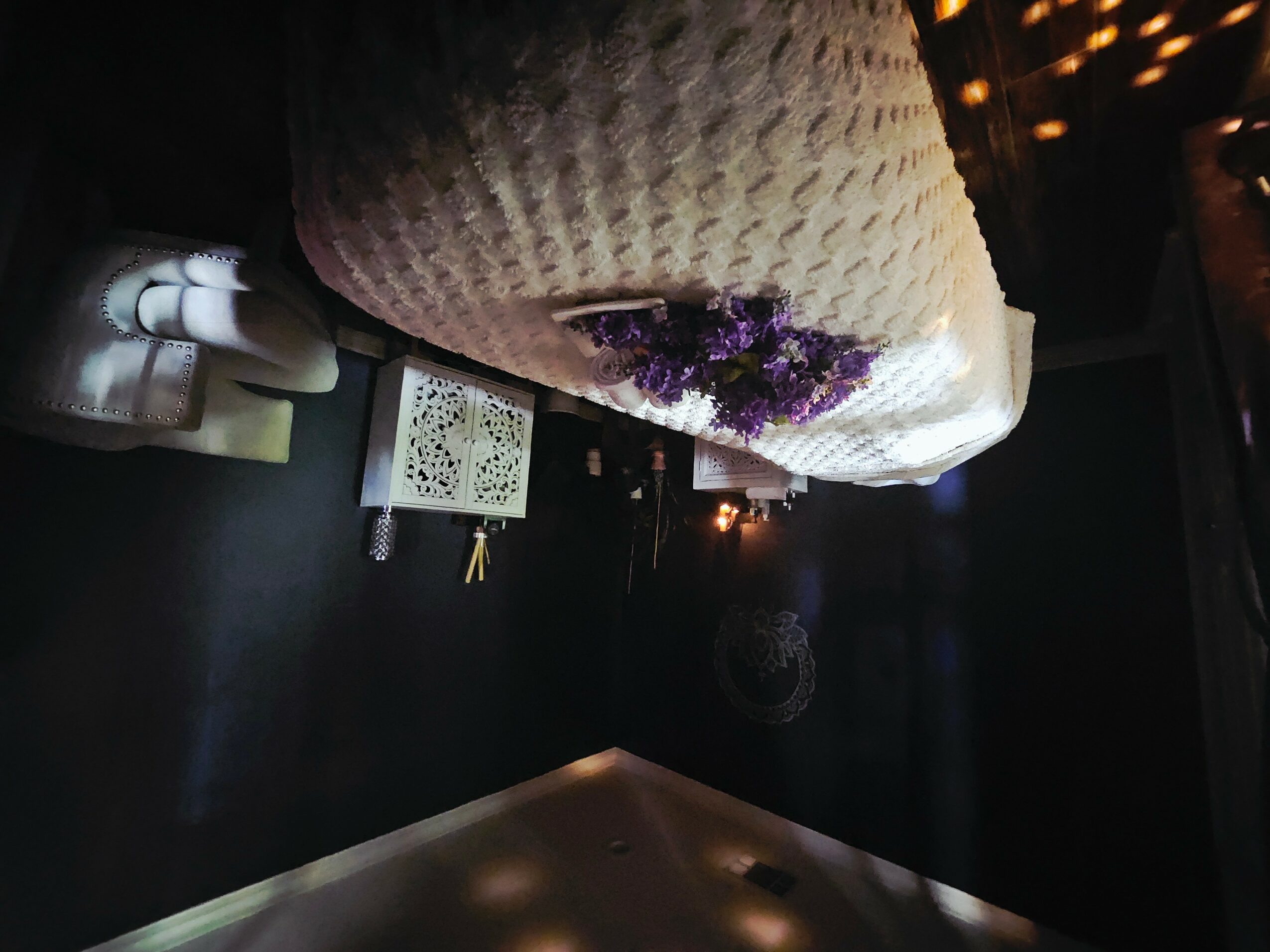 A bed with a pillow and purple flowers, a nightstand with bottles, and a dark wall with a small light.
