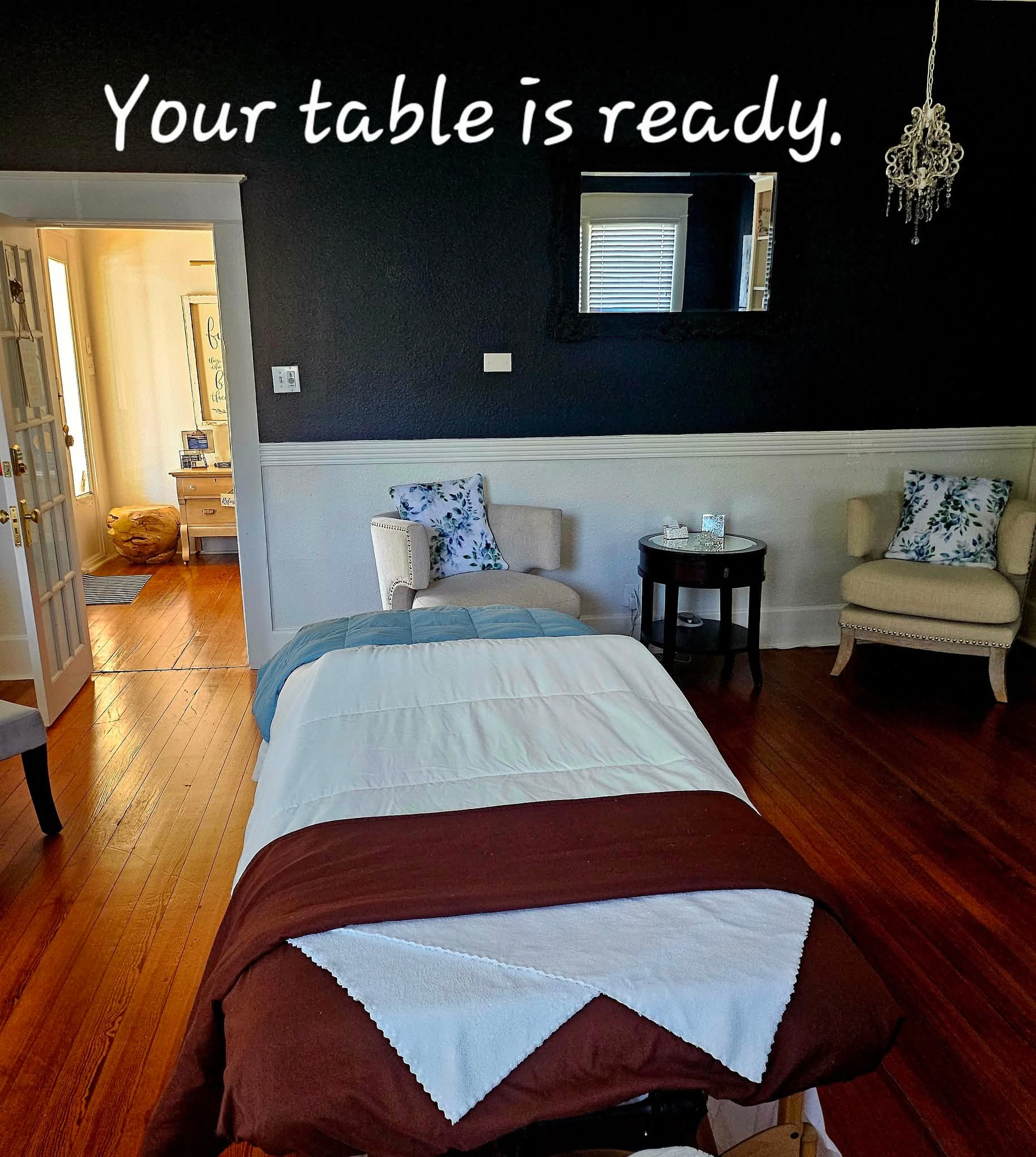 Bedroom with bed, two chairs, small table, dark wall, chandelier, and window, ready for guests.