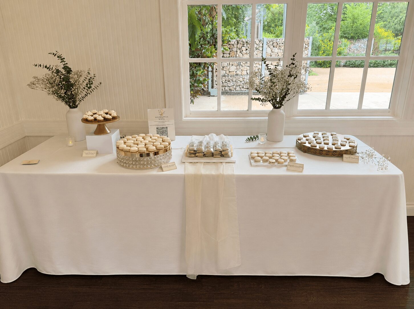 Table with desserts and decorations, window with greenery outside in background, white tablecloth, vases with flowers, desserts on plates