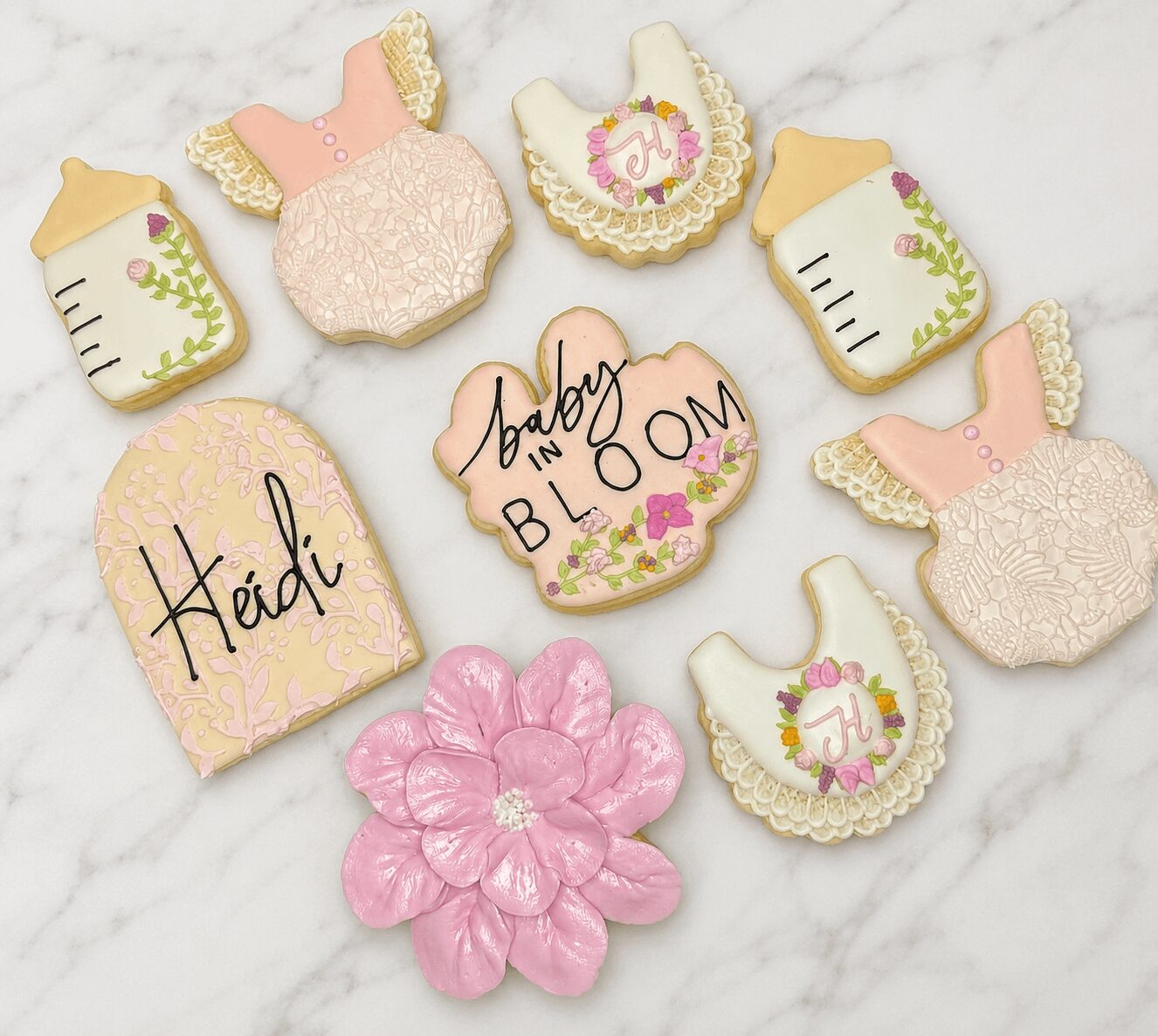 Decorative cookies with baby shower themes, including bottles, dresses, a flower, and personalized names Heidi and Baby Bloom, on a marble surface.