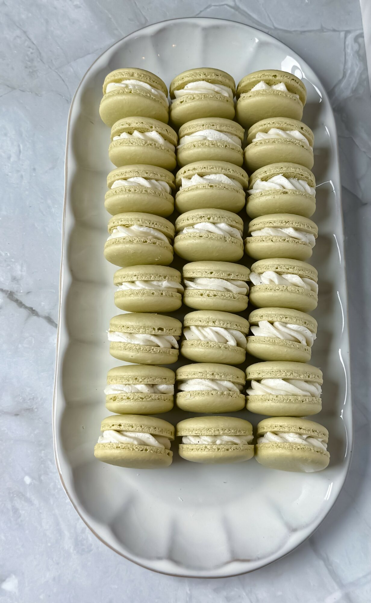 A white oval plate with 24 yellow macarons arranged in four rows on a marble surface.