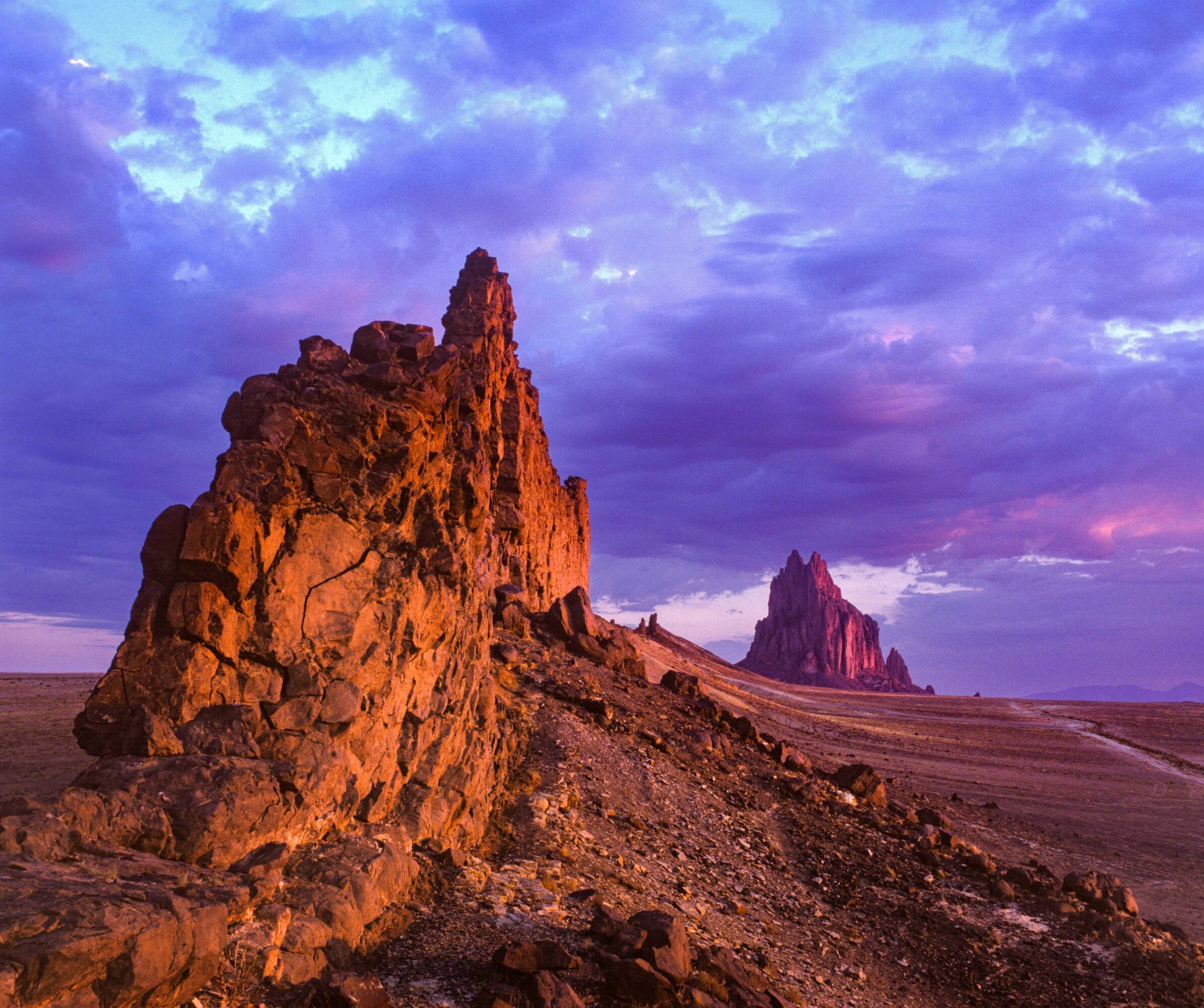 Rock formations in a desert landscape under a colorful sky with clouds, purple, pink, and blue hues.