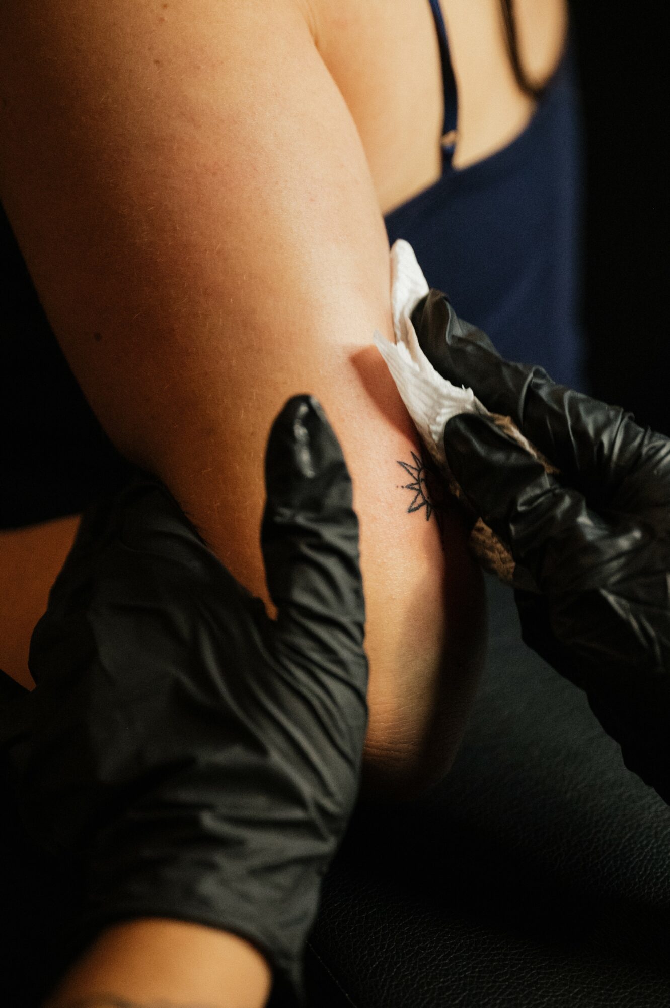 Person receiving a tattoo on their arm, with a tattoo artist wearing black gloves cleaning the area.