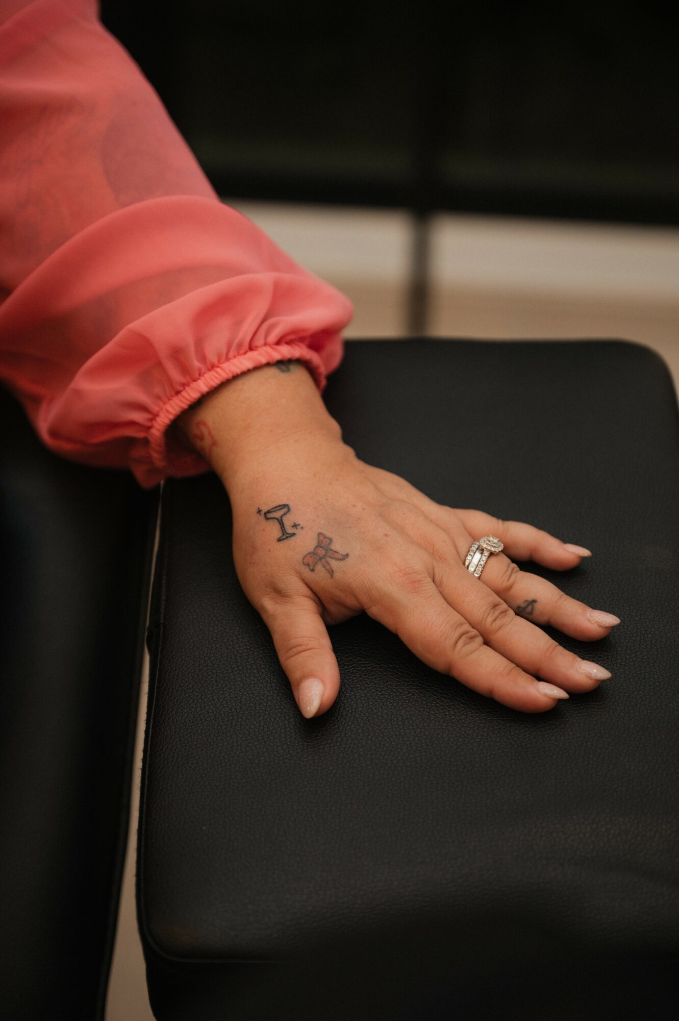 Hand resting on a black surface, wearing a pink sleeve, with tattoos and rings visible.