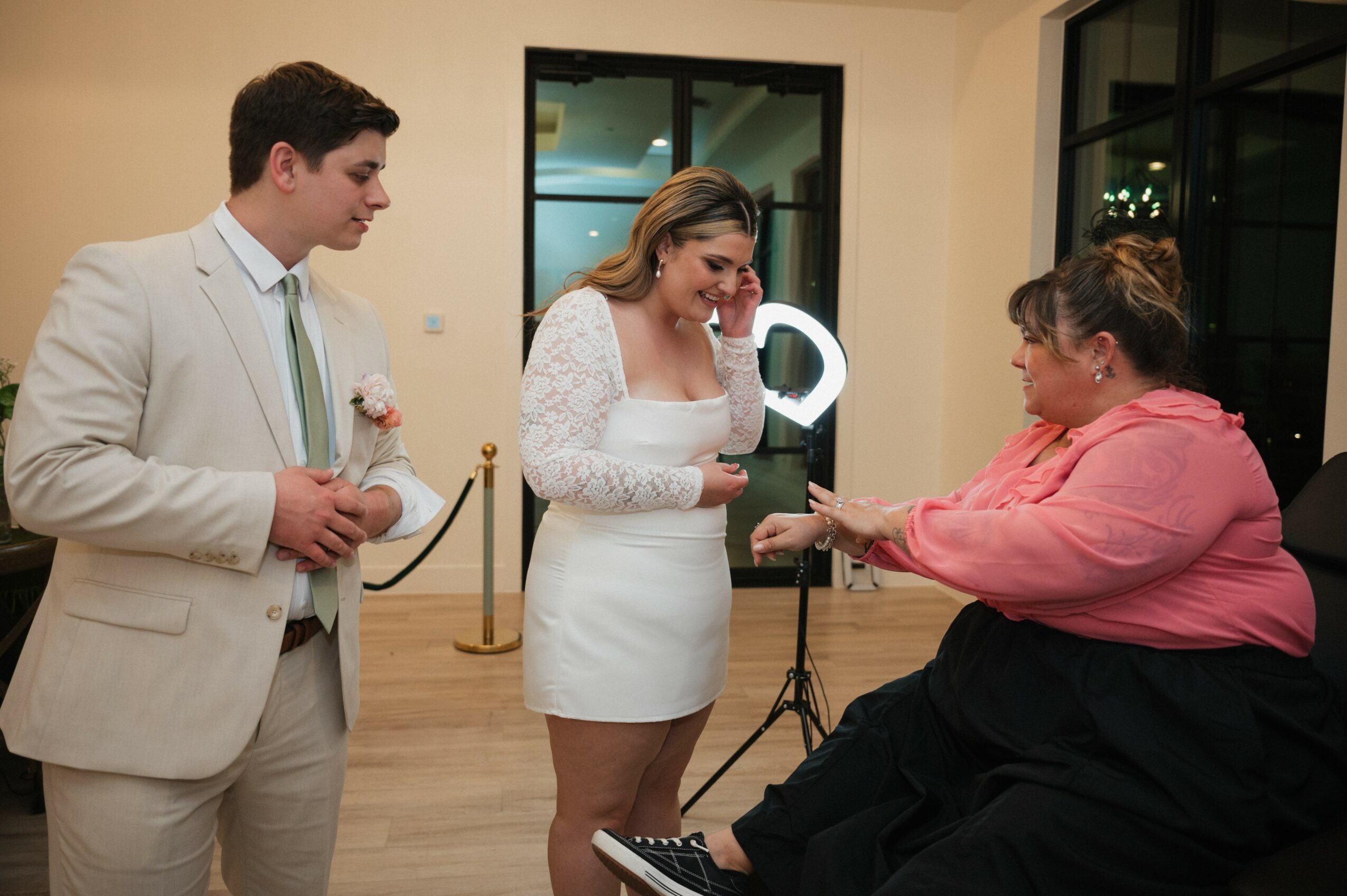 Three people in a room, one woman in a white dress receiving a ring from a woman in a pink top, man in a beige suit watching.