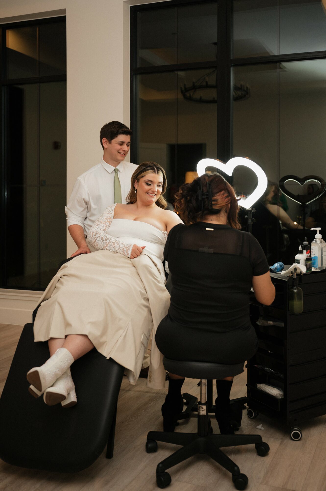 A woman in a beige dress sits on a black chair, getting makeup applied by a makeup artist, with a man standing nearby.