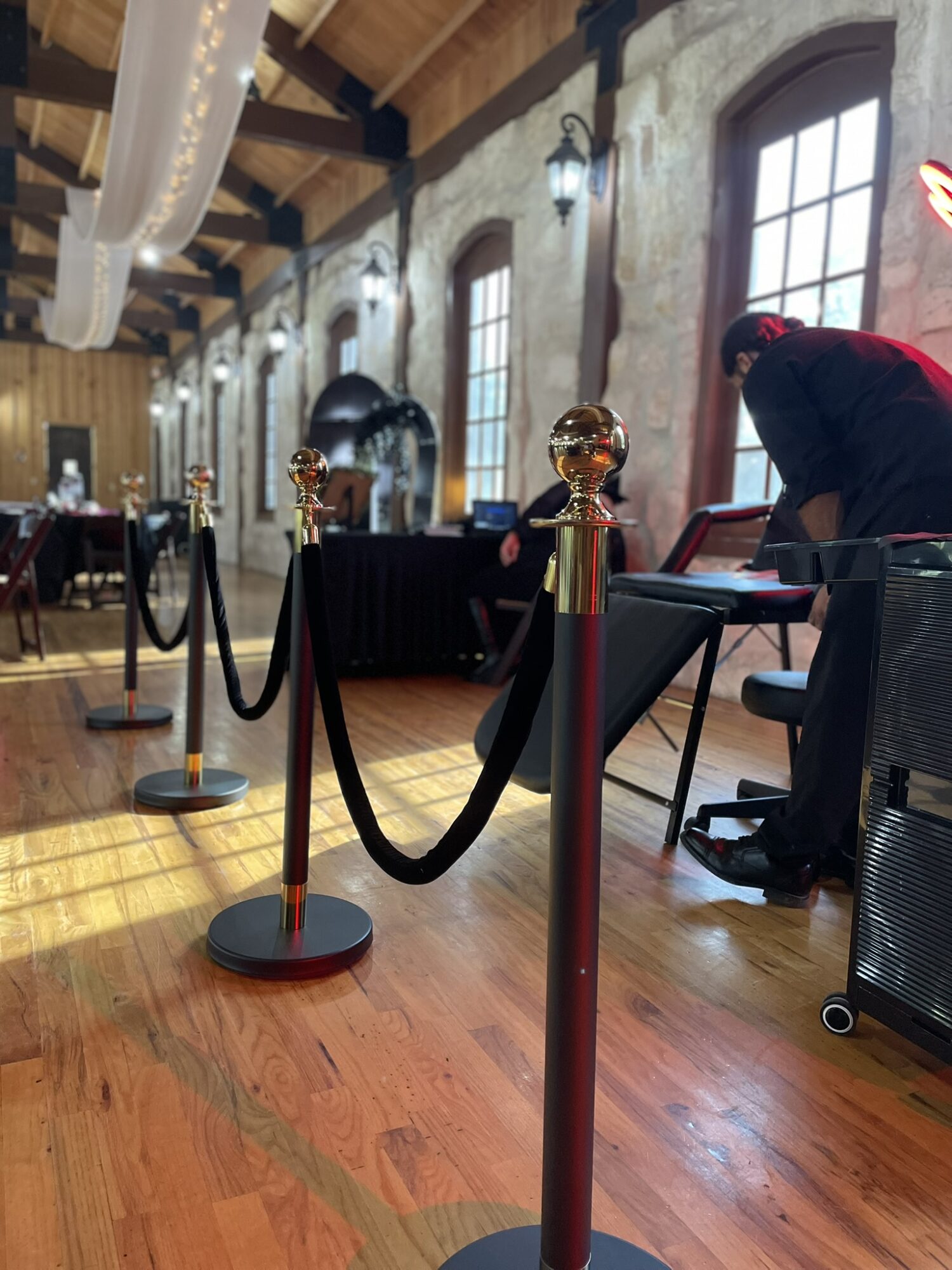 Interior of a rustic event space with wooden floors, brick walls, large windows, and black tables with gold decorations.