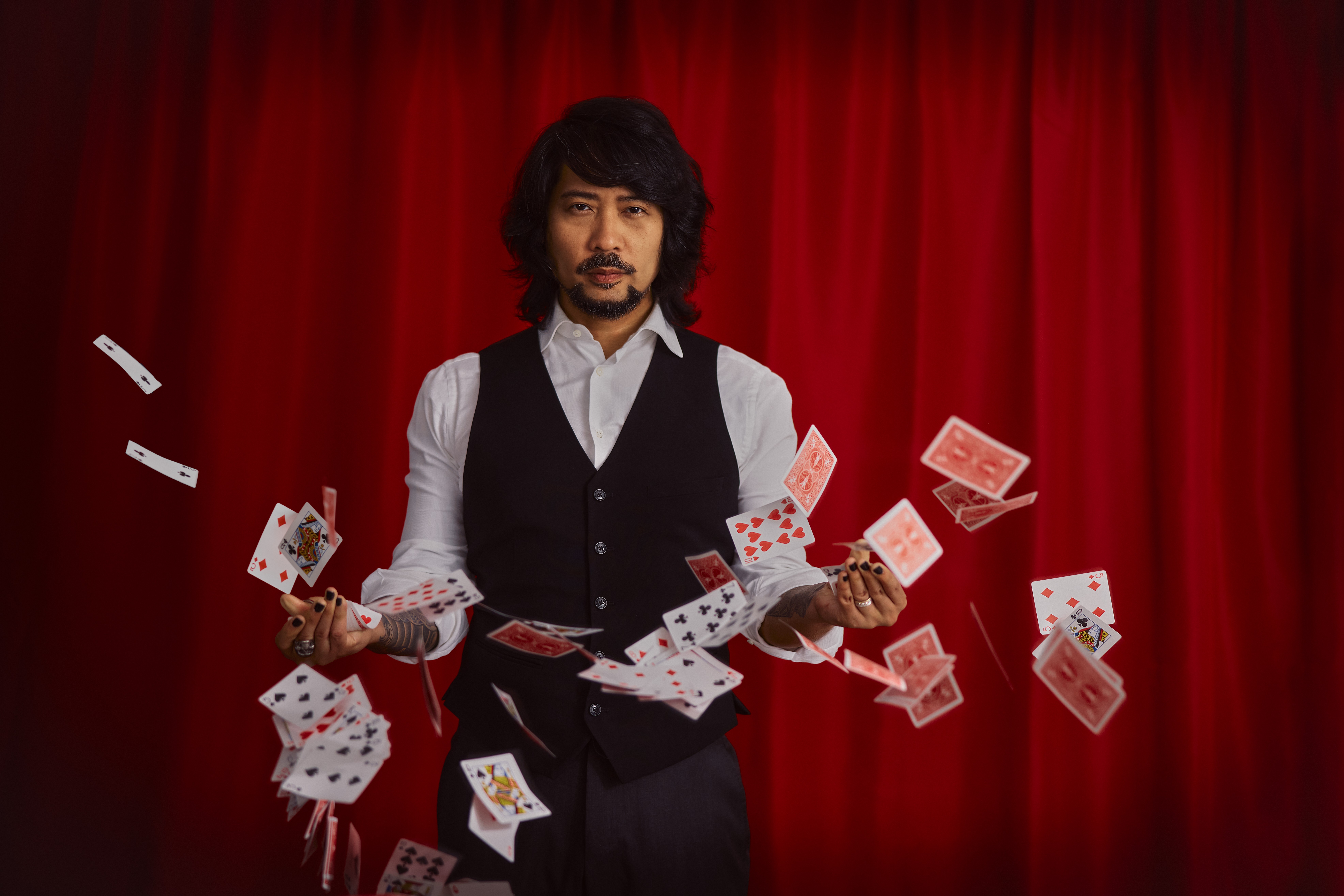 Man performing card trick with playing cards in front of red curtain, cards flying around him.