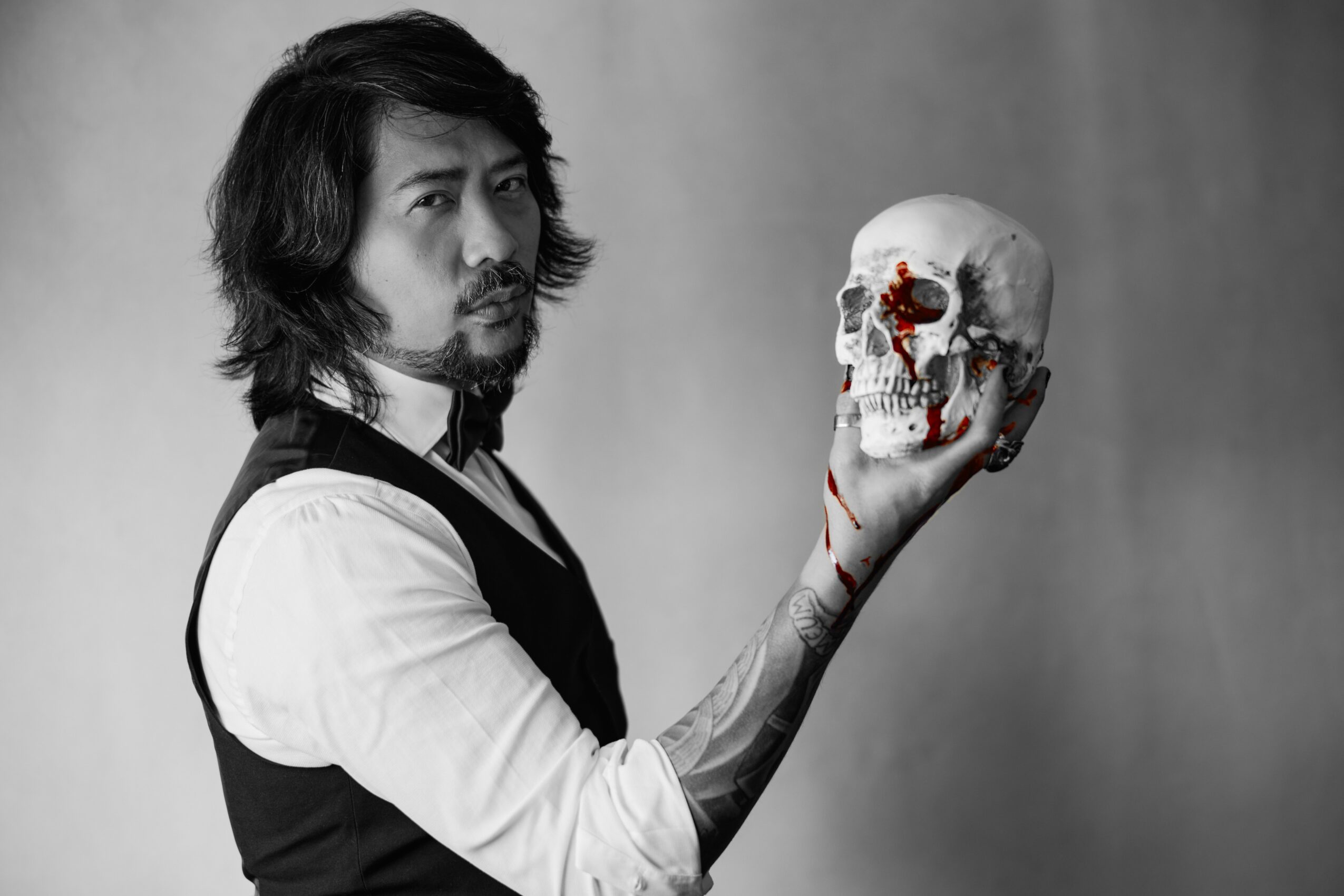 Man with long hair and beard holding a skull with red eye socket, black and white background.