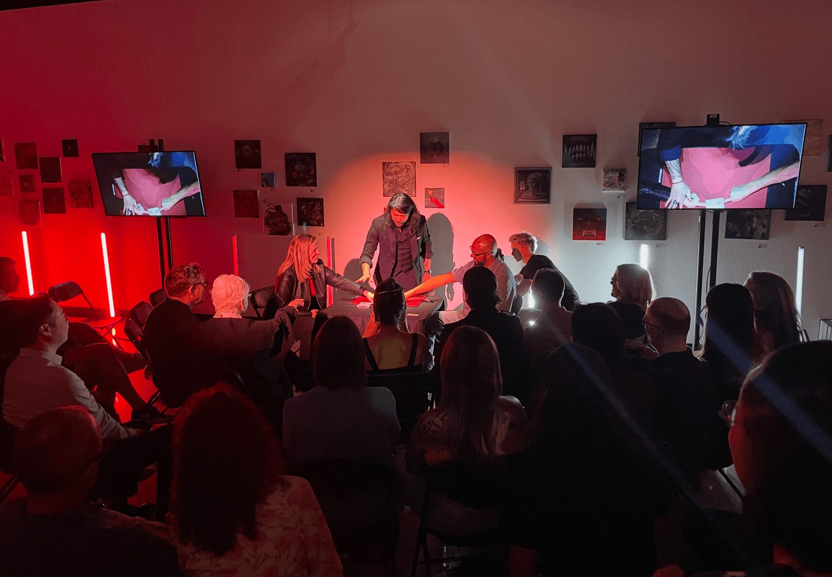 Person performing on stage with audience watching, two screens displaying visuals, red lighting, and wall decorations in background.