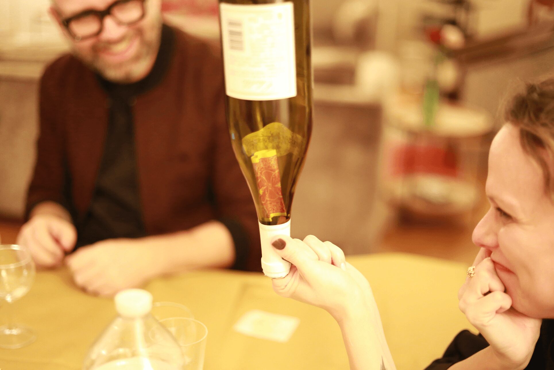Person holding a bottle upside down over a glass, with another person smiling nearby, in a cozy indoor setting.