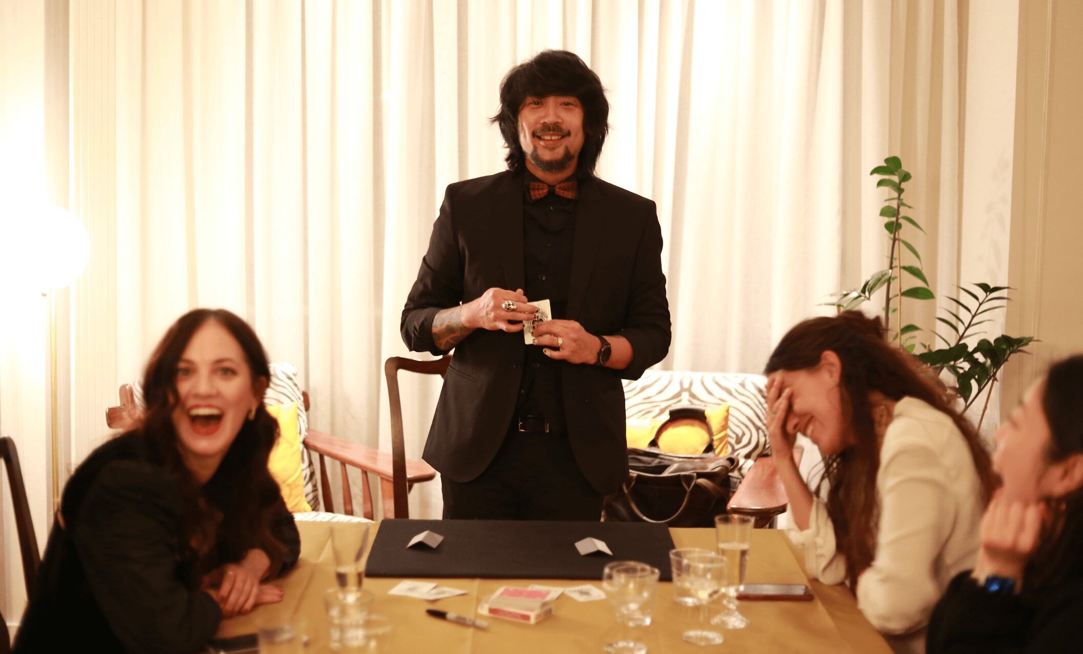 Four people sitting around a table, one standing, smiling and laughing, in a room with curtains and plants.