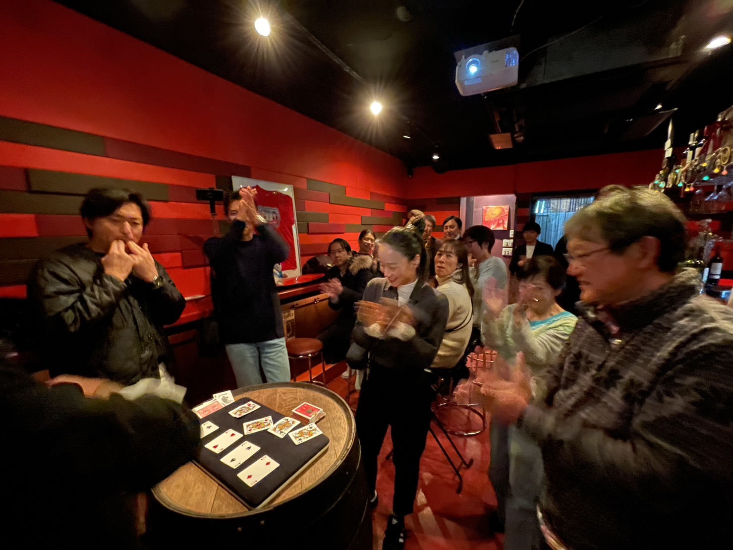 People gathered around a table playing cards in a dimly lit room with red walls and a bar in the background.