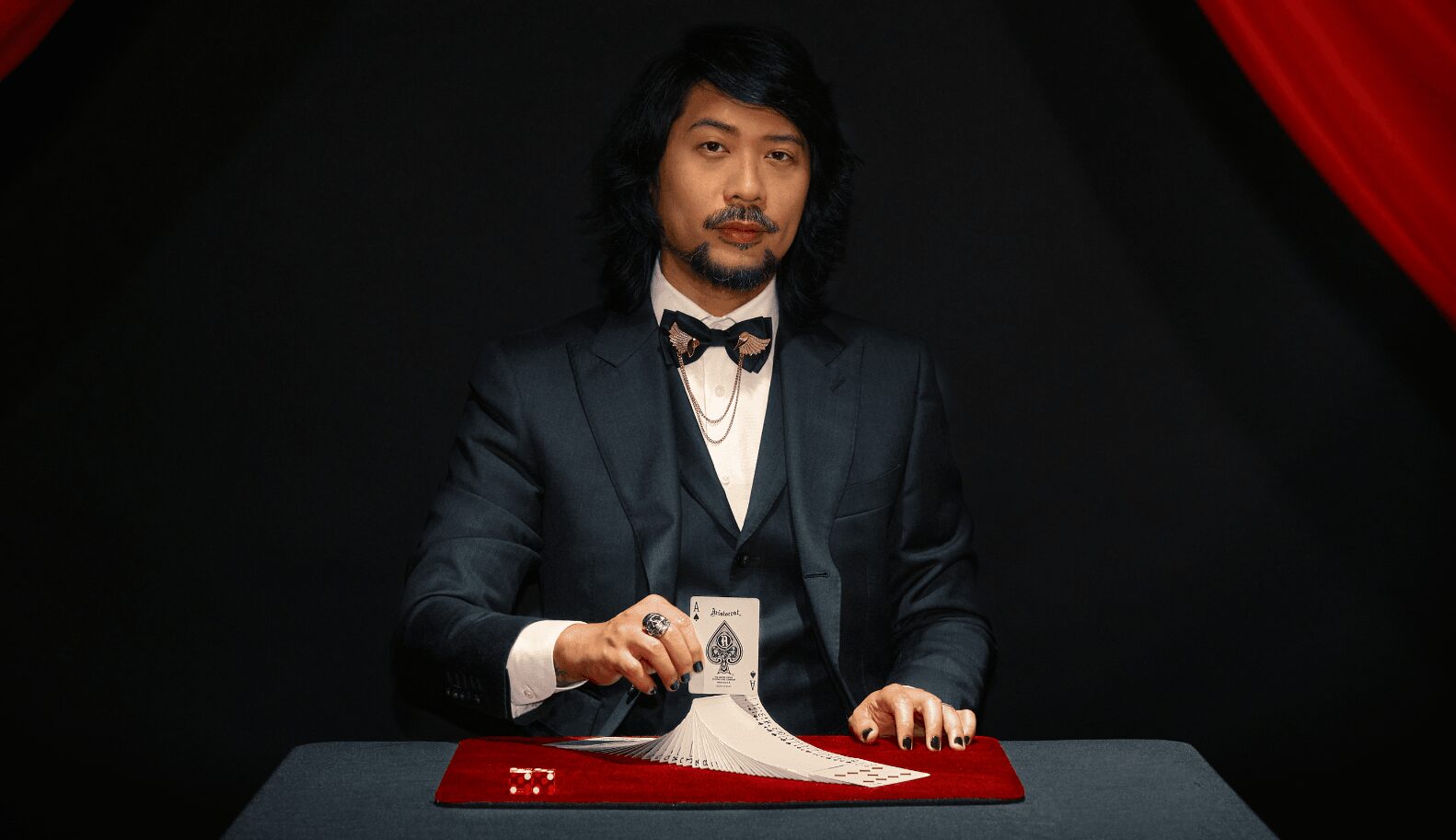 Man in formal suit and bow tie sitting at a table with a red cloth, holding a pen, against a dark background with red curtains.