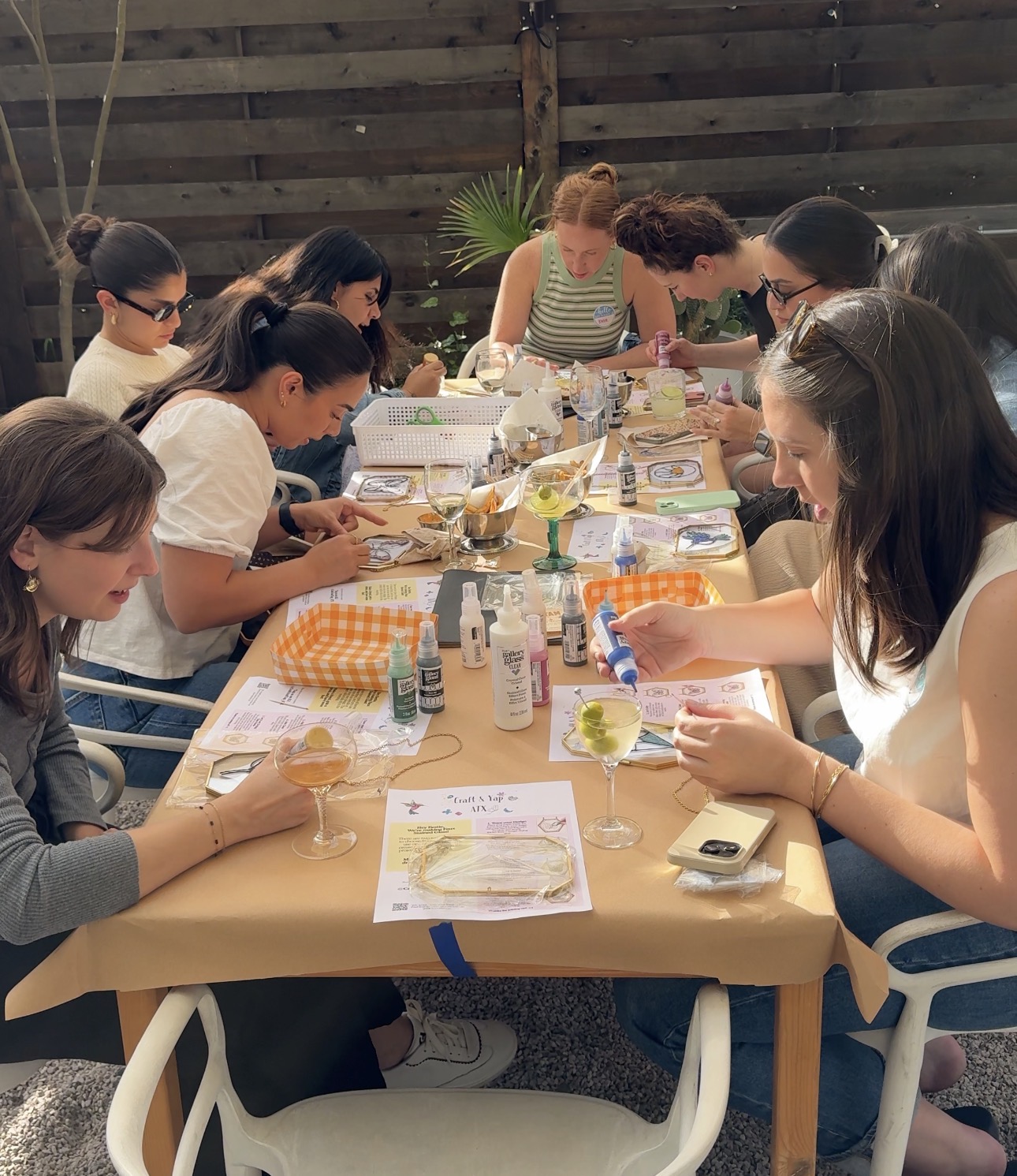 Group of women around a table making crafts outdoors, with various supplies and a wooden fence in background.