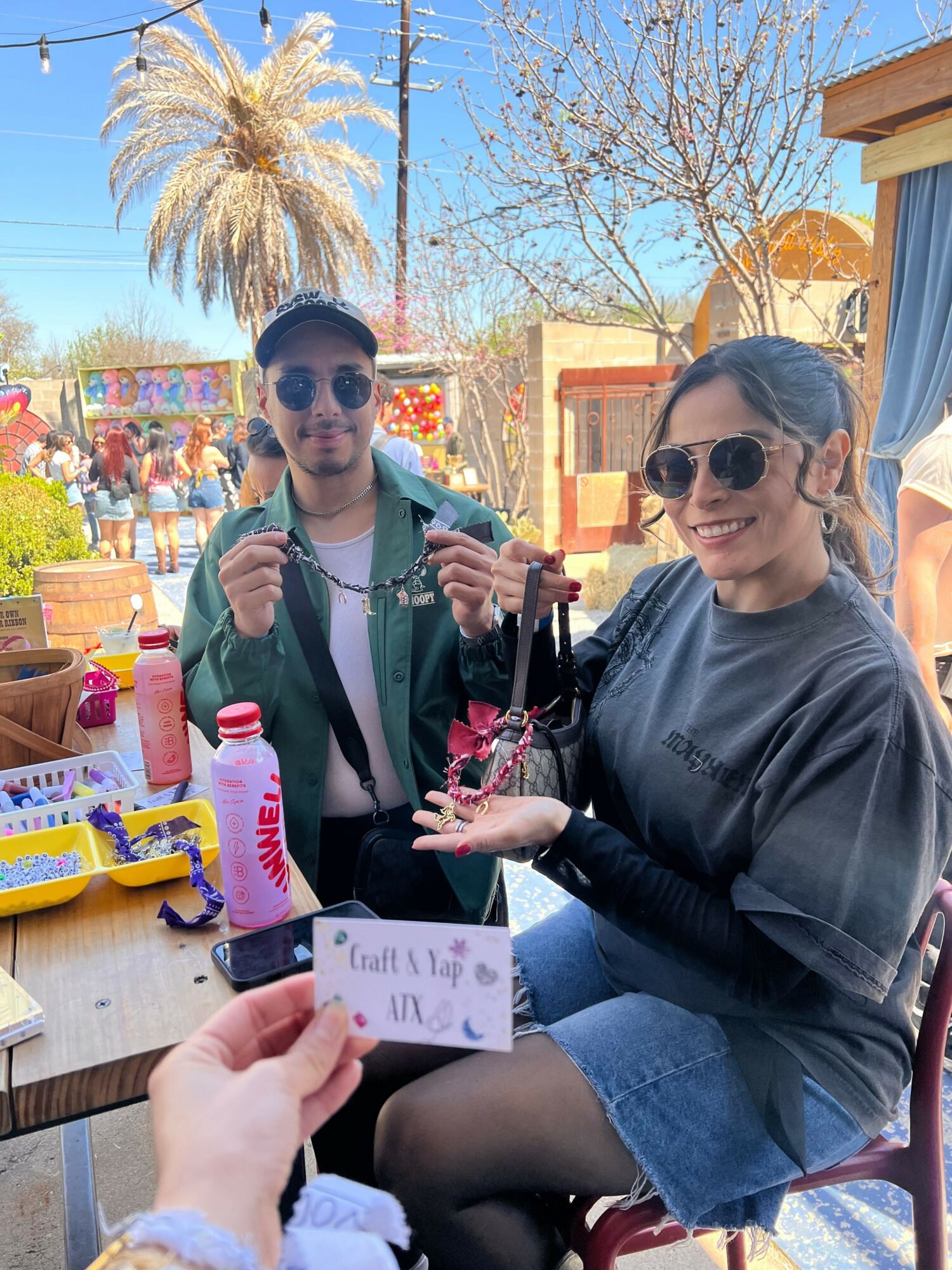 Two women sitting outdoors at a market or fair, one holding a card, with colorful booths and trees in background.
