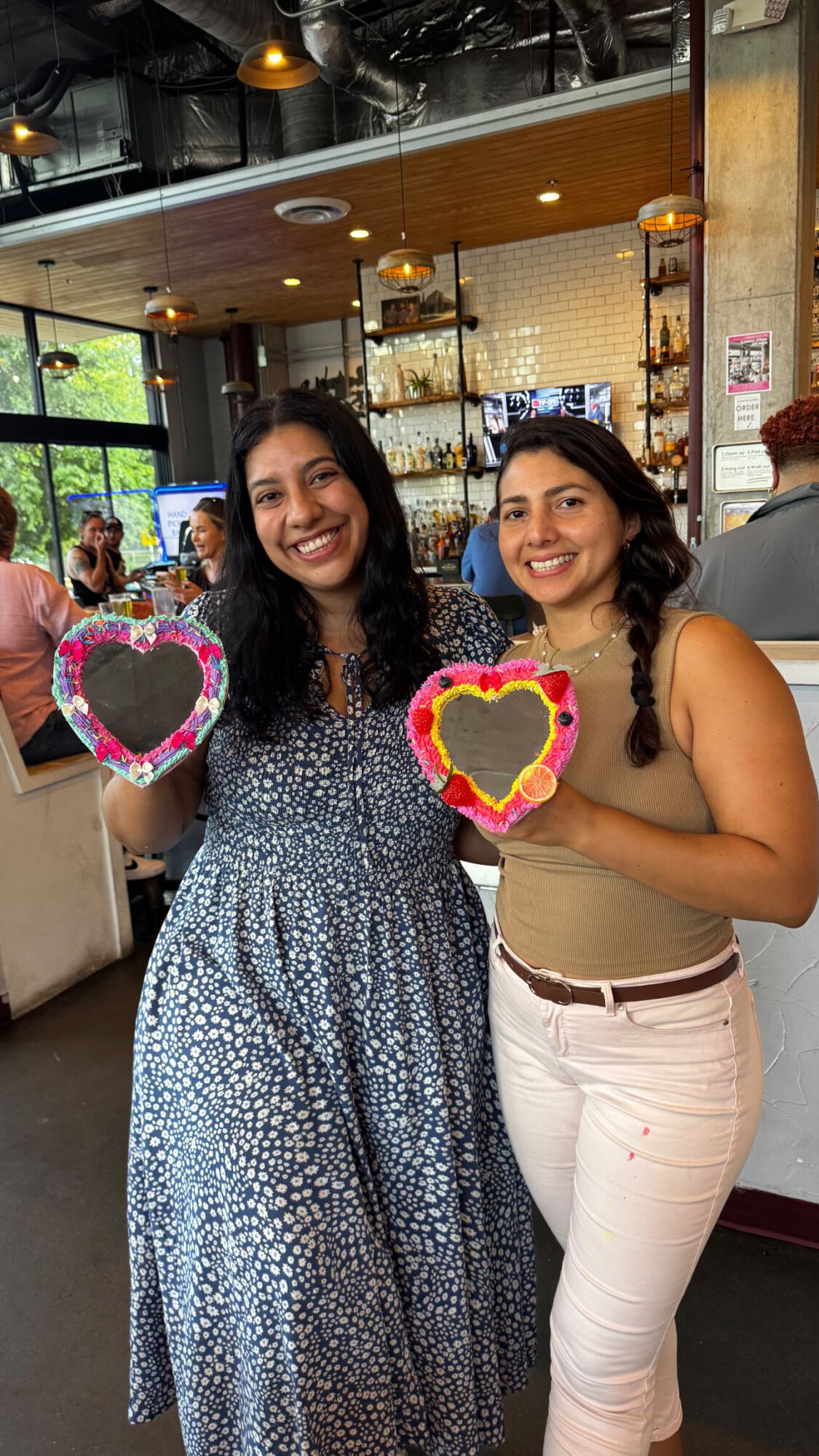 Two women smiling indoors, each holding a heart-shaped decoration with colorful borders, in a lively setting with people and shelves in background.
