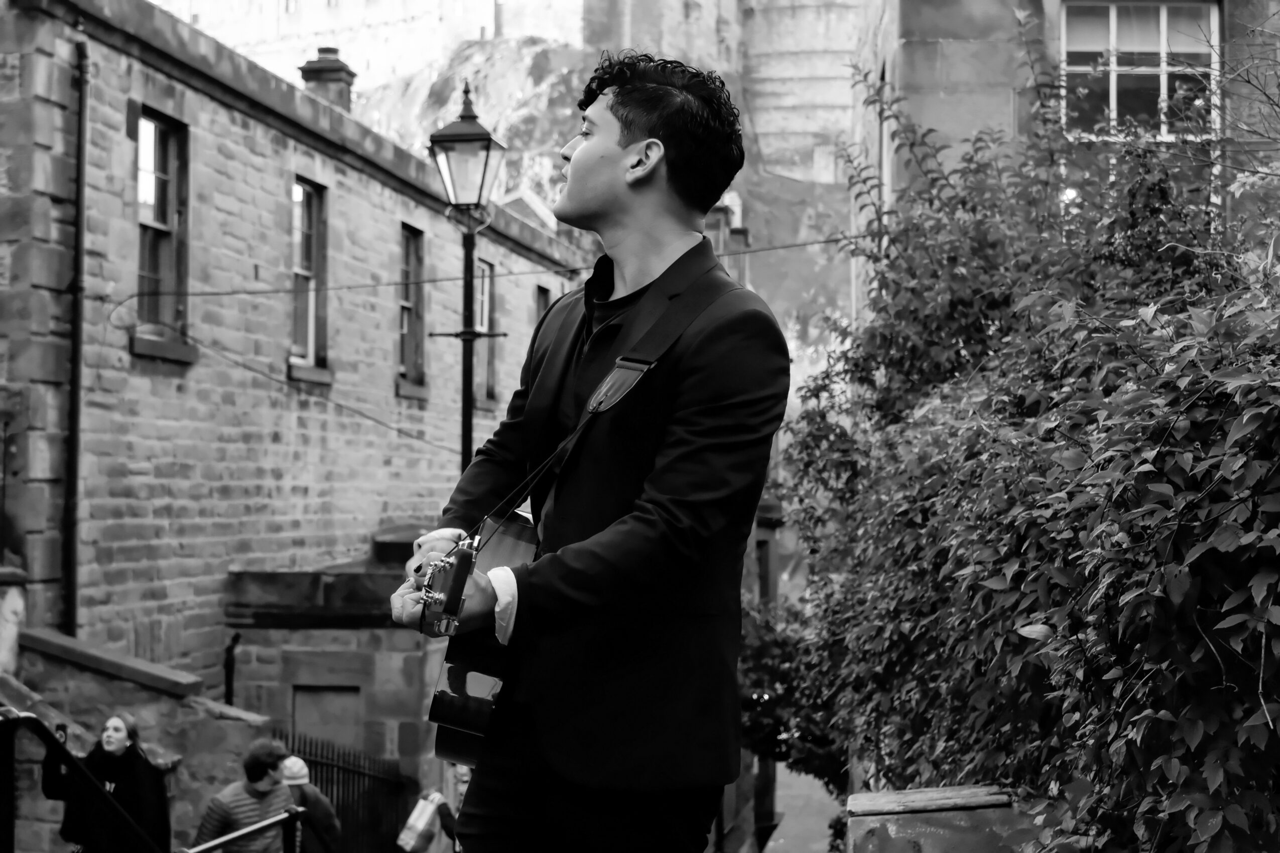 Man in a suit standing outdoors near a brick building and bushes, looking to the side.
