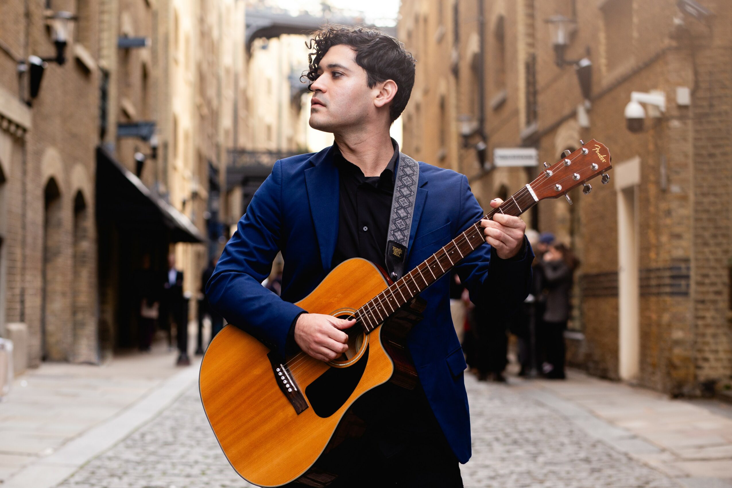 Young man with dark hair playing an acoustic guitar in a narrow brick alleyway, wearing a blue jacket and black shirt.