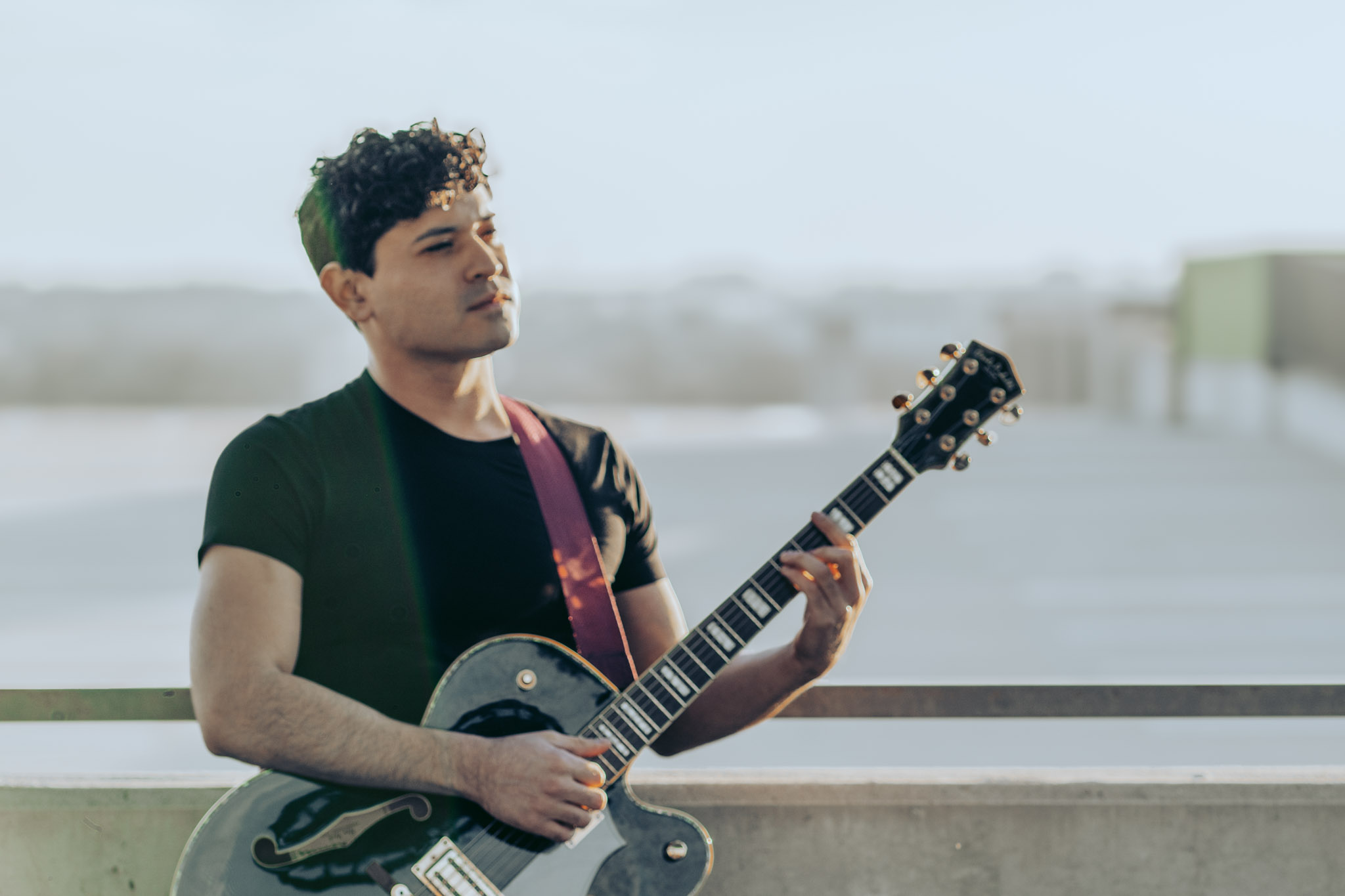 Young man with curly hair playing a guitar outdoors, standing near a railing with a blurred background.