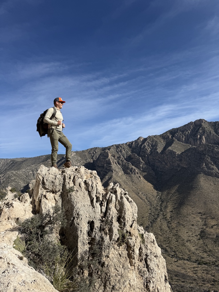 Person standing on a large rock formation in a mountainous landscape under a blue sky with wispy clouds.