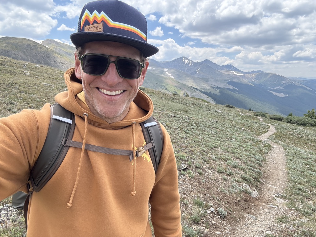 Smiling man wearing sunglasses, a cap, and a brown hoodie, hiking on a mountain trail with scenic landscape.